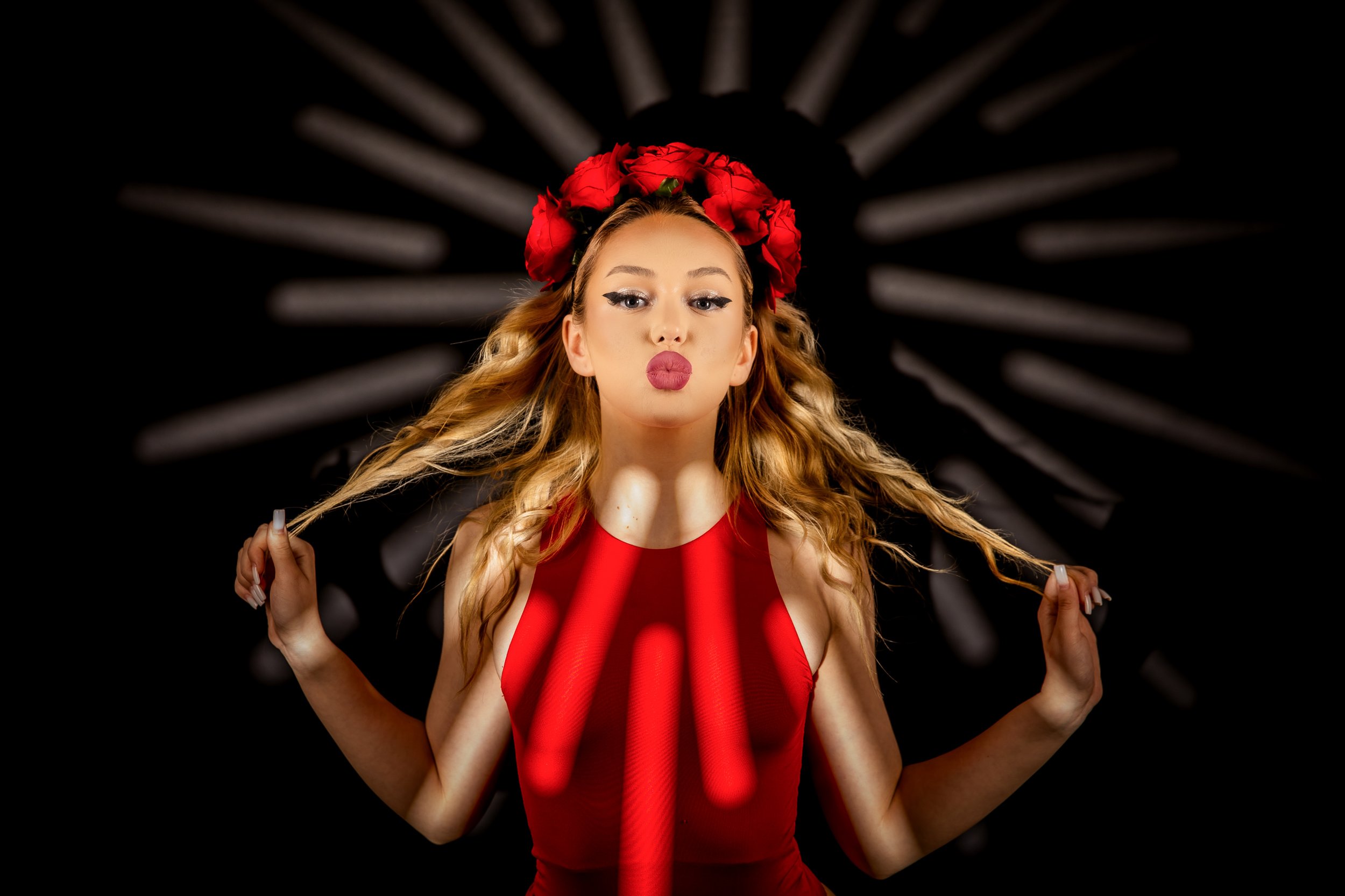 A woman with long wavy blonde hair wears a red dress and a red flower crown, puckering her lips with a blurred background of black with white radial patterns.