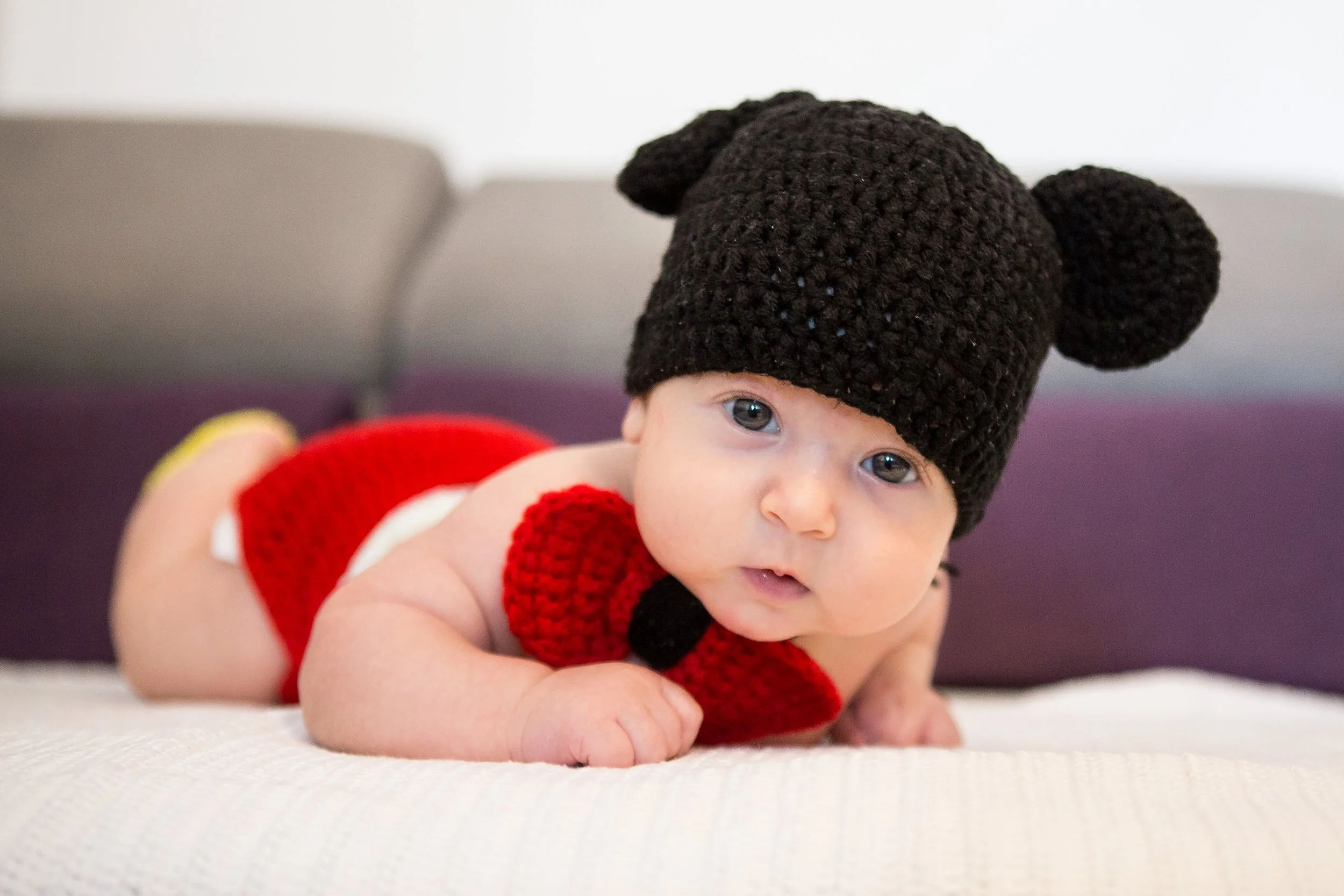 A baby crawling on a white textured surface wearing a red crochet outfit and a black crochet hat with bear ears.