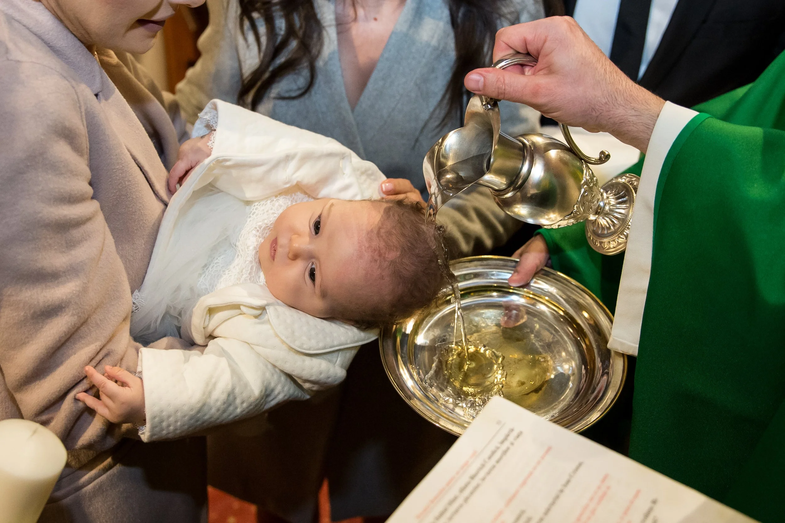 A young child lies down on an adult's arm during a baptism ceremony, with an officiant pouring water over the child's head from a silver baptismal vessel.