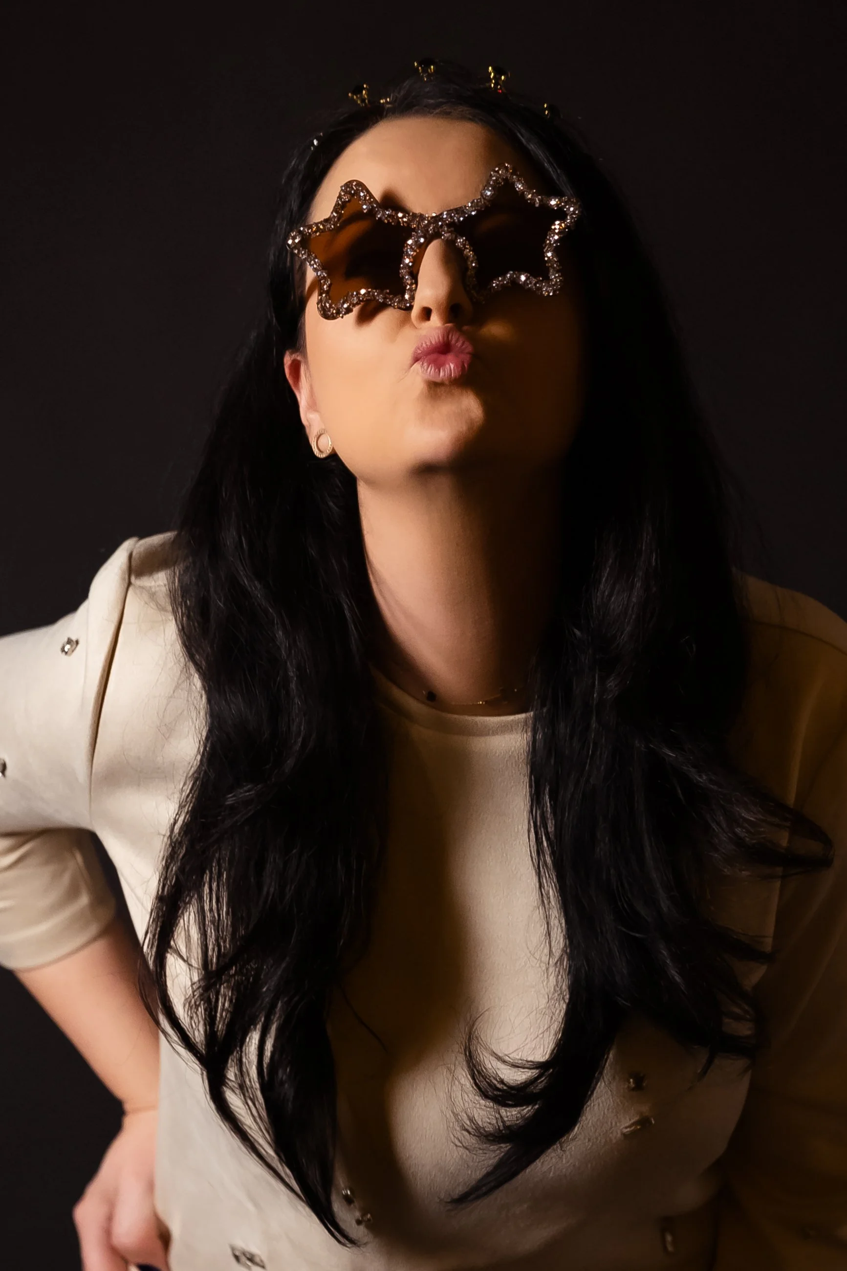 A woman with long black hair wearing star-shaped sunglasses with rhinestones, a beige jacket with metallic buttons, and small hoop earrings, against a black background.