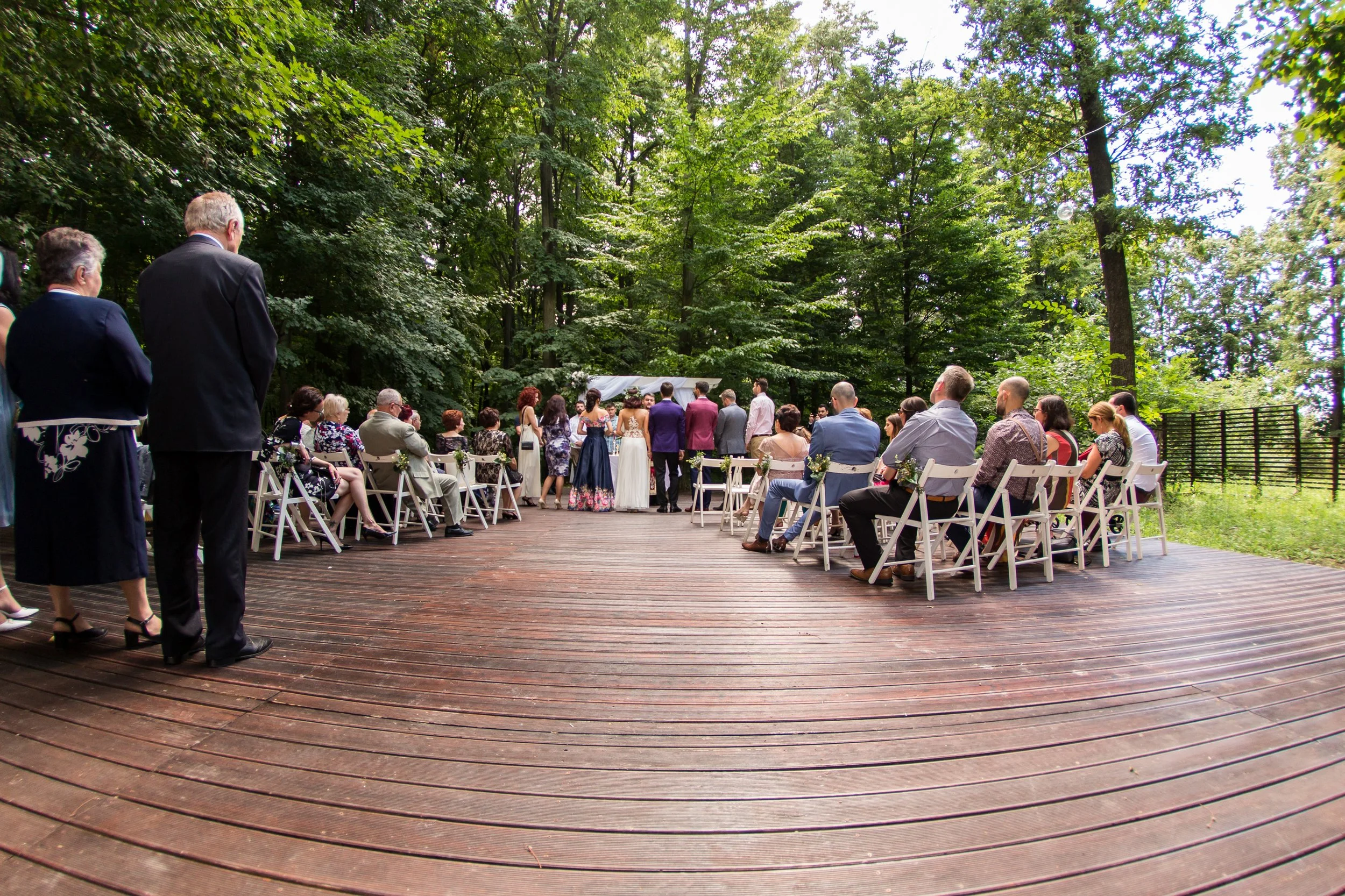 People attending an outdoor wedding ceremony in a wooded area with chairs arranged in rows and a white arch at the front.
