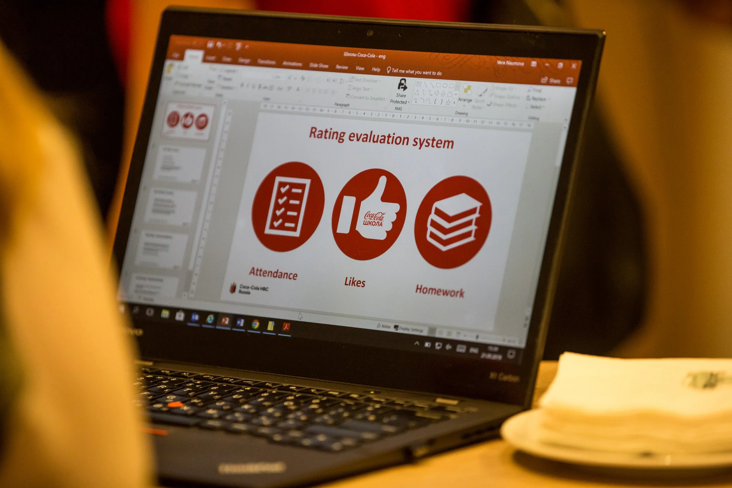 A laptop on a wooden desk displaying a PowerPoint slide titled 'Rating evaluation system' with icons for attendance, likes, and homework.
