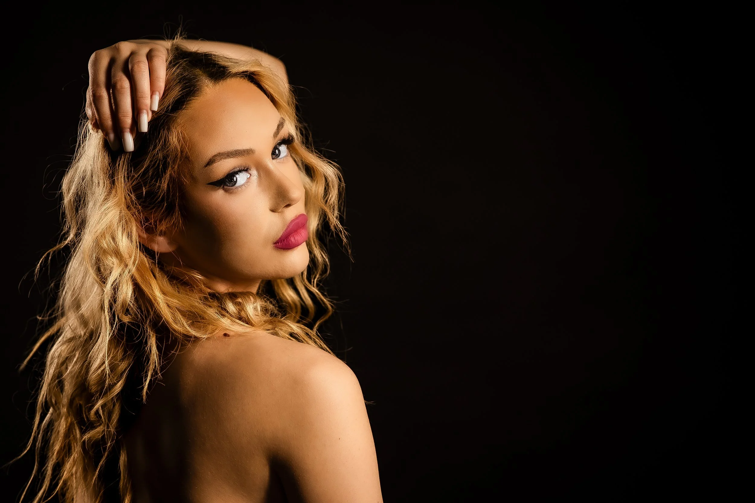 A woman with long, curly blonde hair and makeup, looking over her shoulder with her hand resting on her head, against a black background.