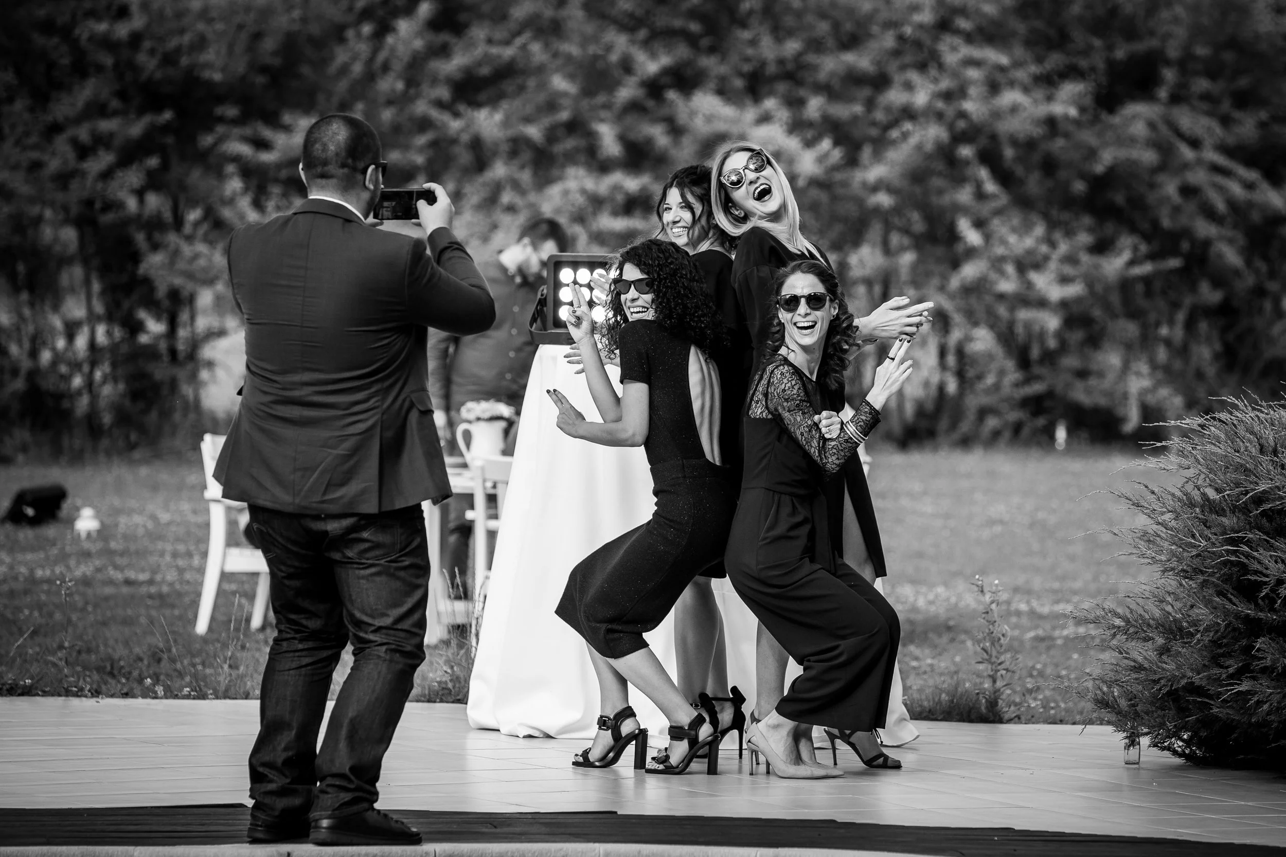 Group of women dancing and taking photos outdoors at a celebration or wedding, with a man in the foreground capturing the moment.