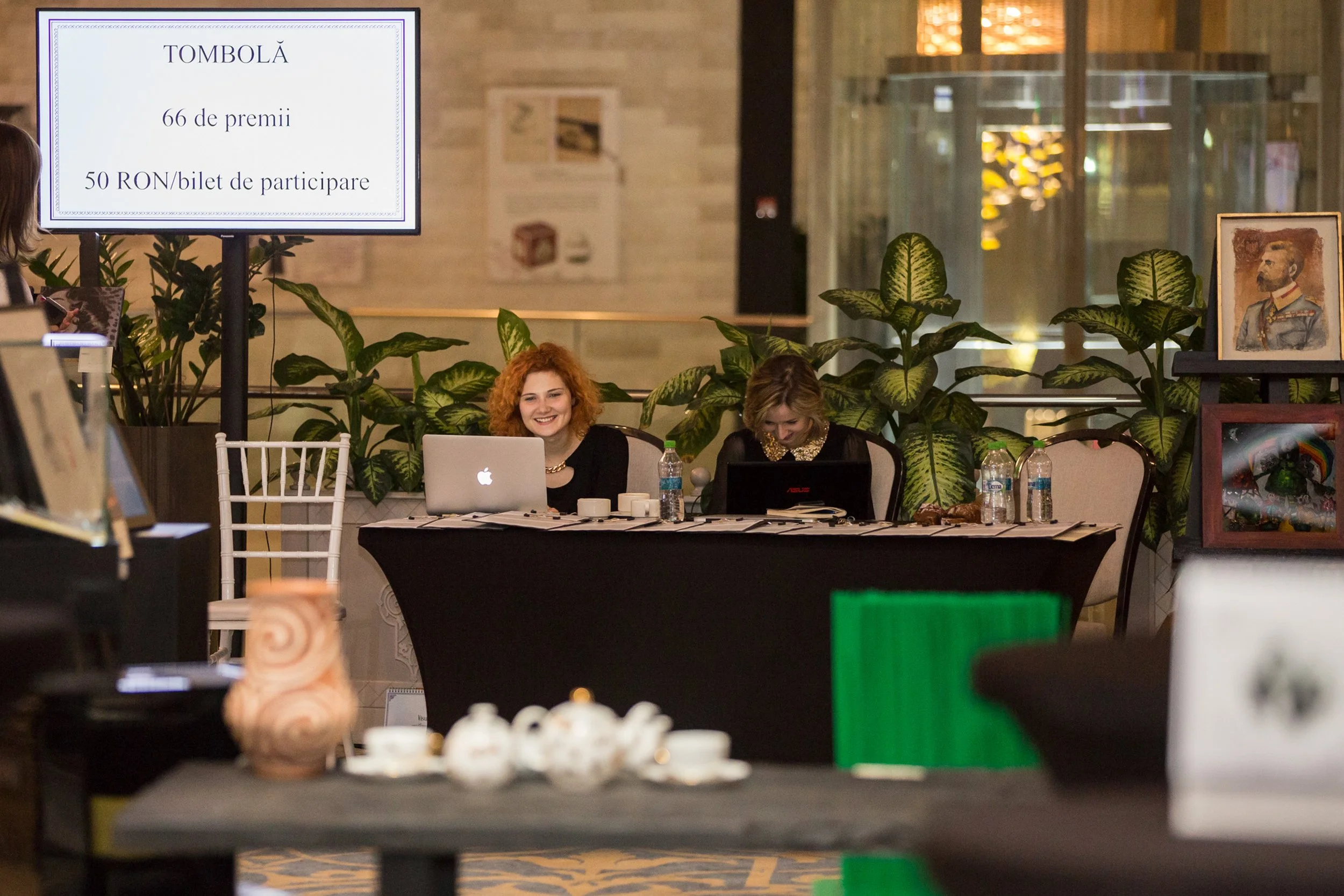 Two women sitting at a table with laptops and a bottle of water, smiling. Behind them are large green plants, framed artwork on display, and a large screen displaying a sign in Romanian about a raffle.