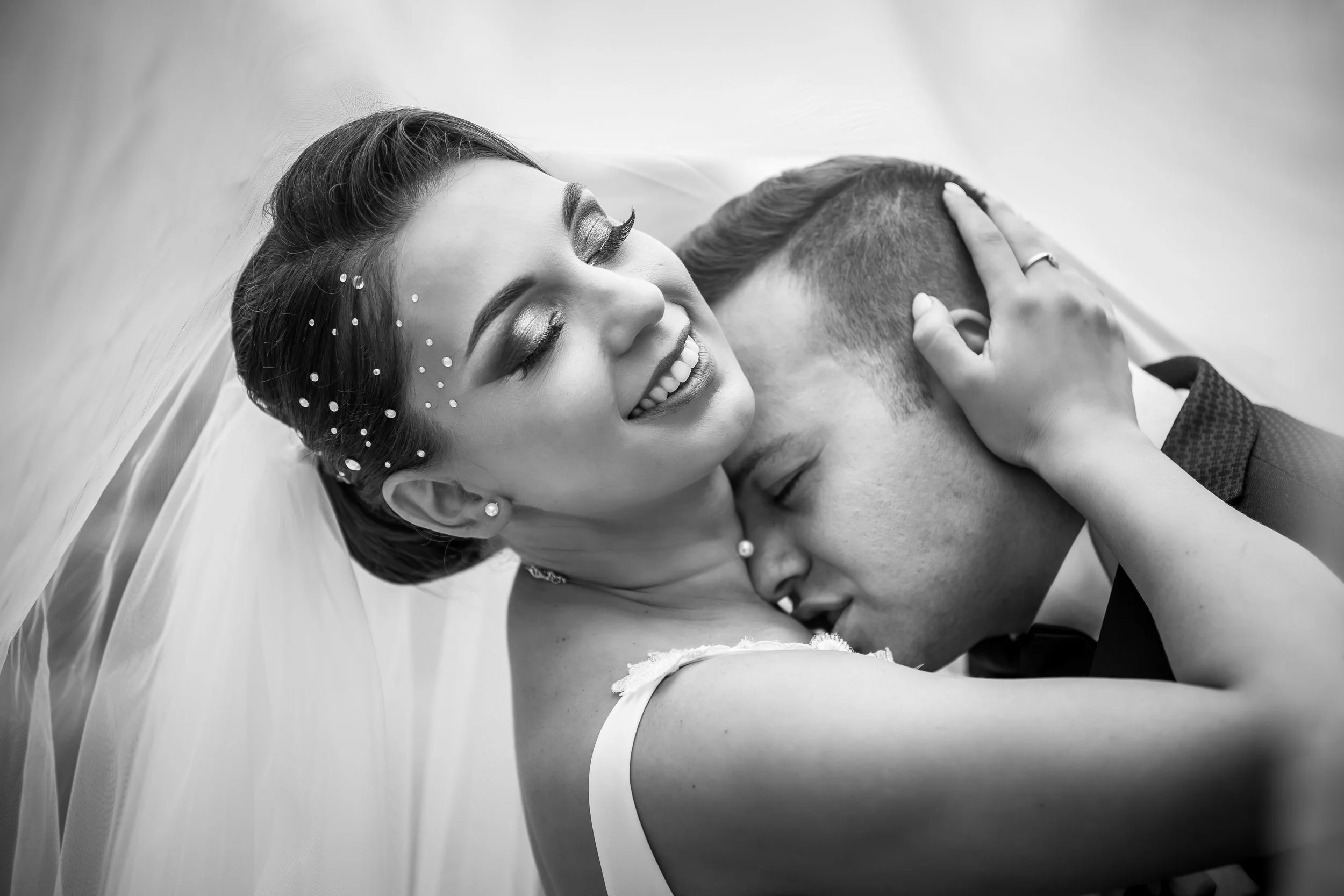 Black and white photo of a bride and groom sharing a kiss, with the bride smiling with her eyes closed, wearing earrings, a veil, and decorative face embellishments, while the groom's face is partially obscured.
