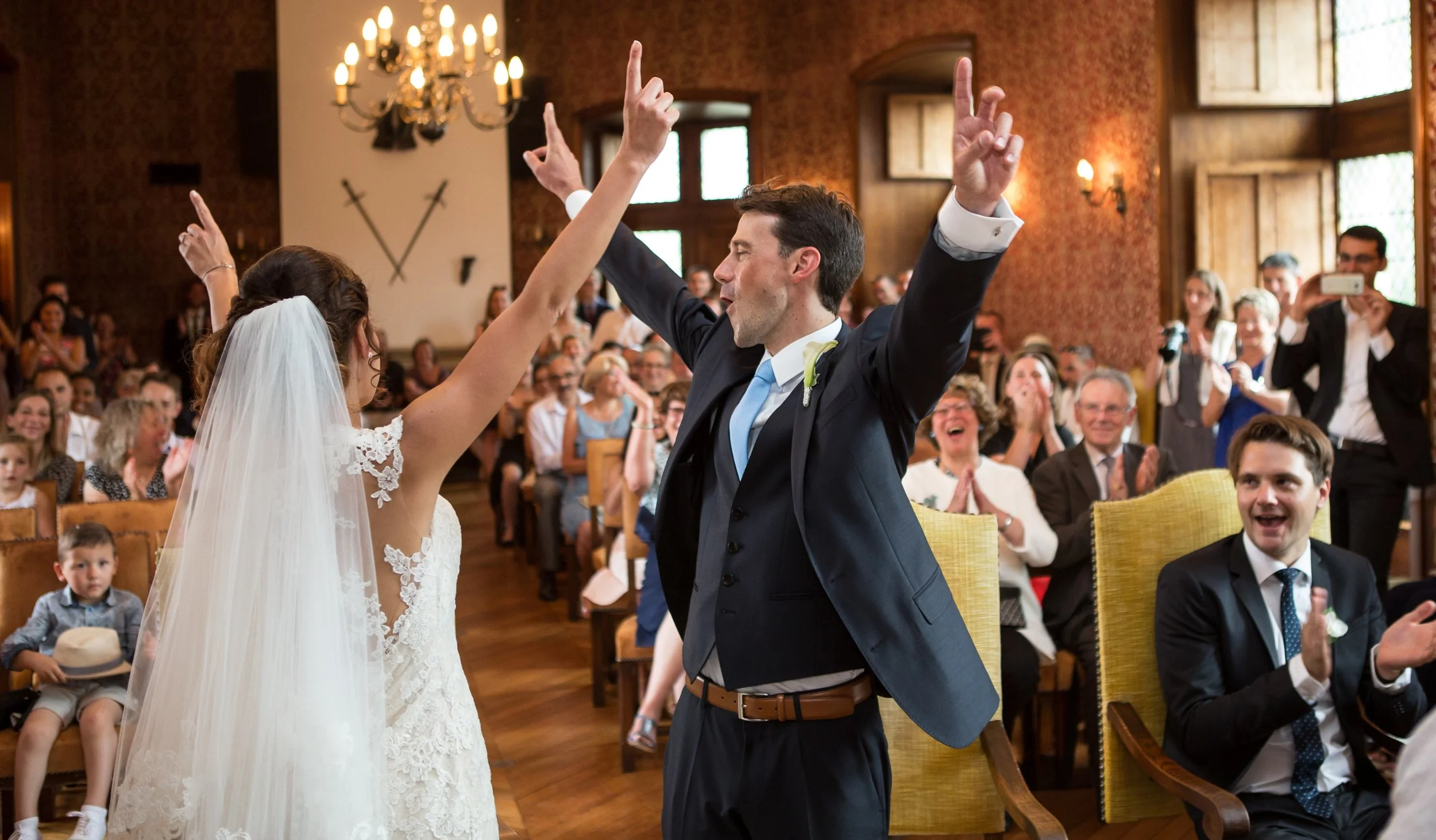 A bride and groom celebrating at their wedding ceremony indoors, waving and smiling, while surrounded by seated guests in a warmly lit room.