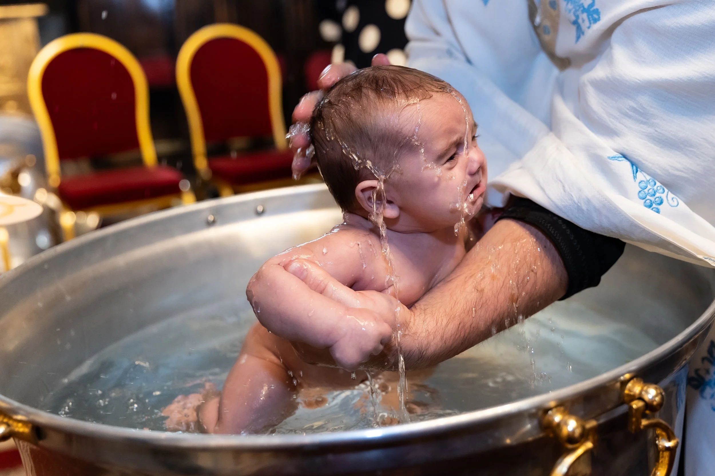 A person holding a baby during a baptism or christening ceremony, with water splashing on the baby's head. The background has red and yellow chairs.