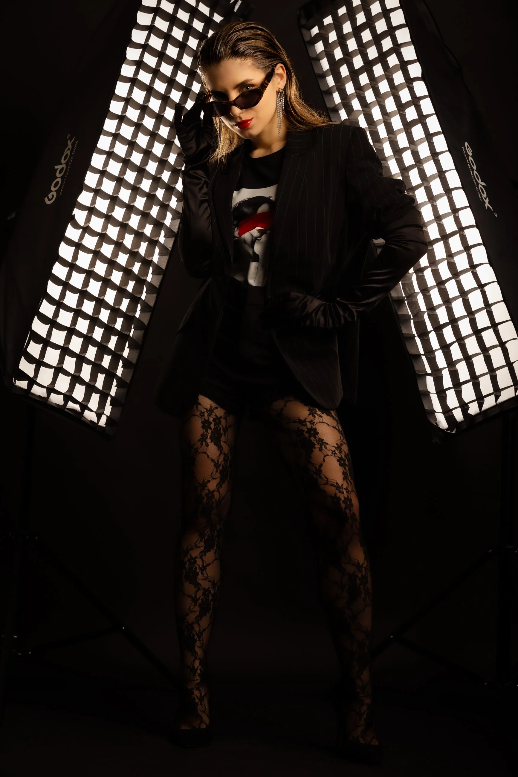 A woman dressed in black, wearing fishnet stockings and sunglasses, standing in front of a professional photography light setup.