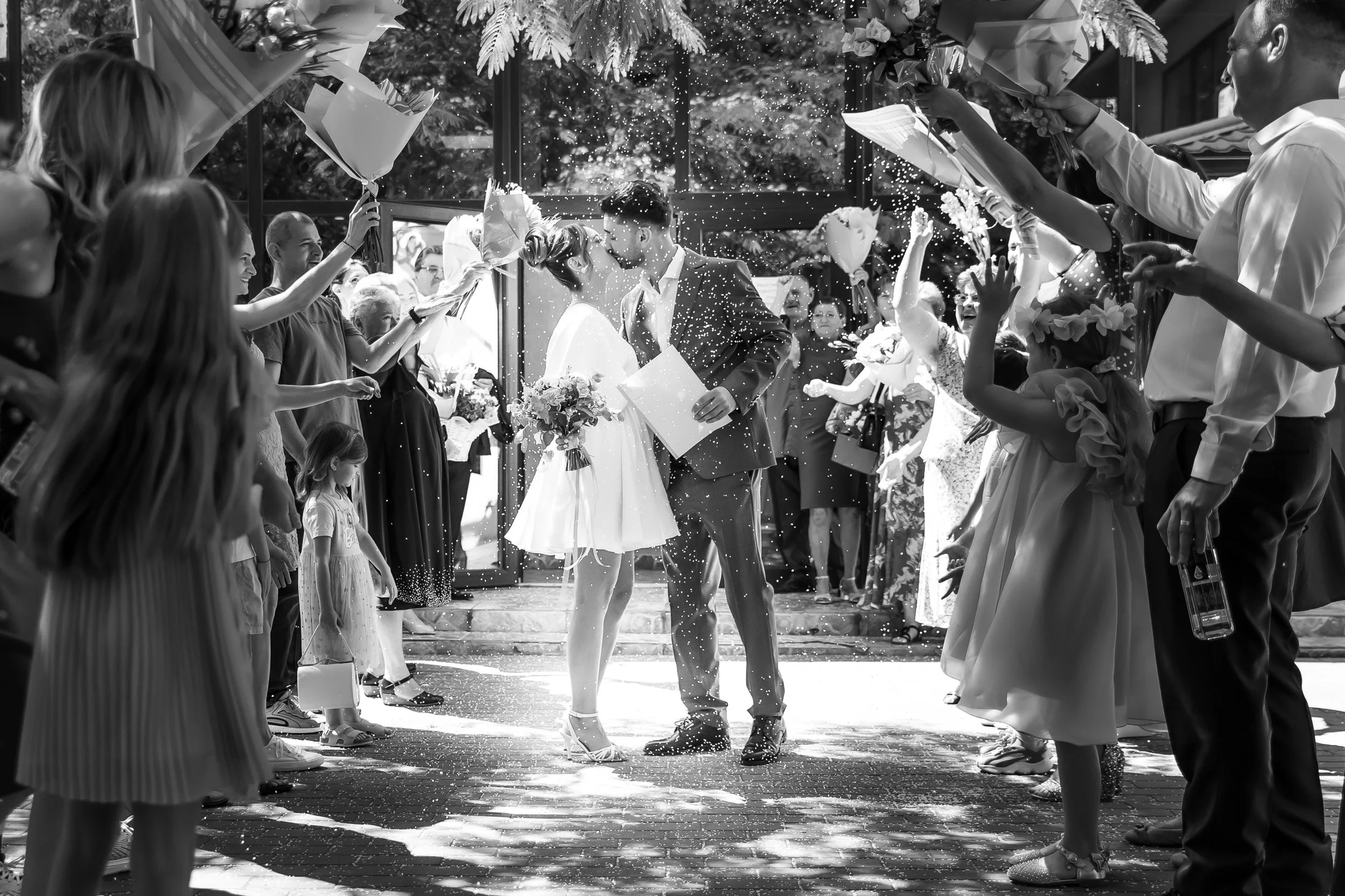 A black and white photo of a wedding celebration with a bride and groom sharing a kiss beside each other while surrounded by friends and family. Guests are throwing confetti and holding bouquets, celebrating outdoors near a glass building with trees 