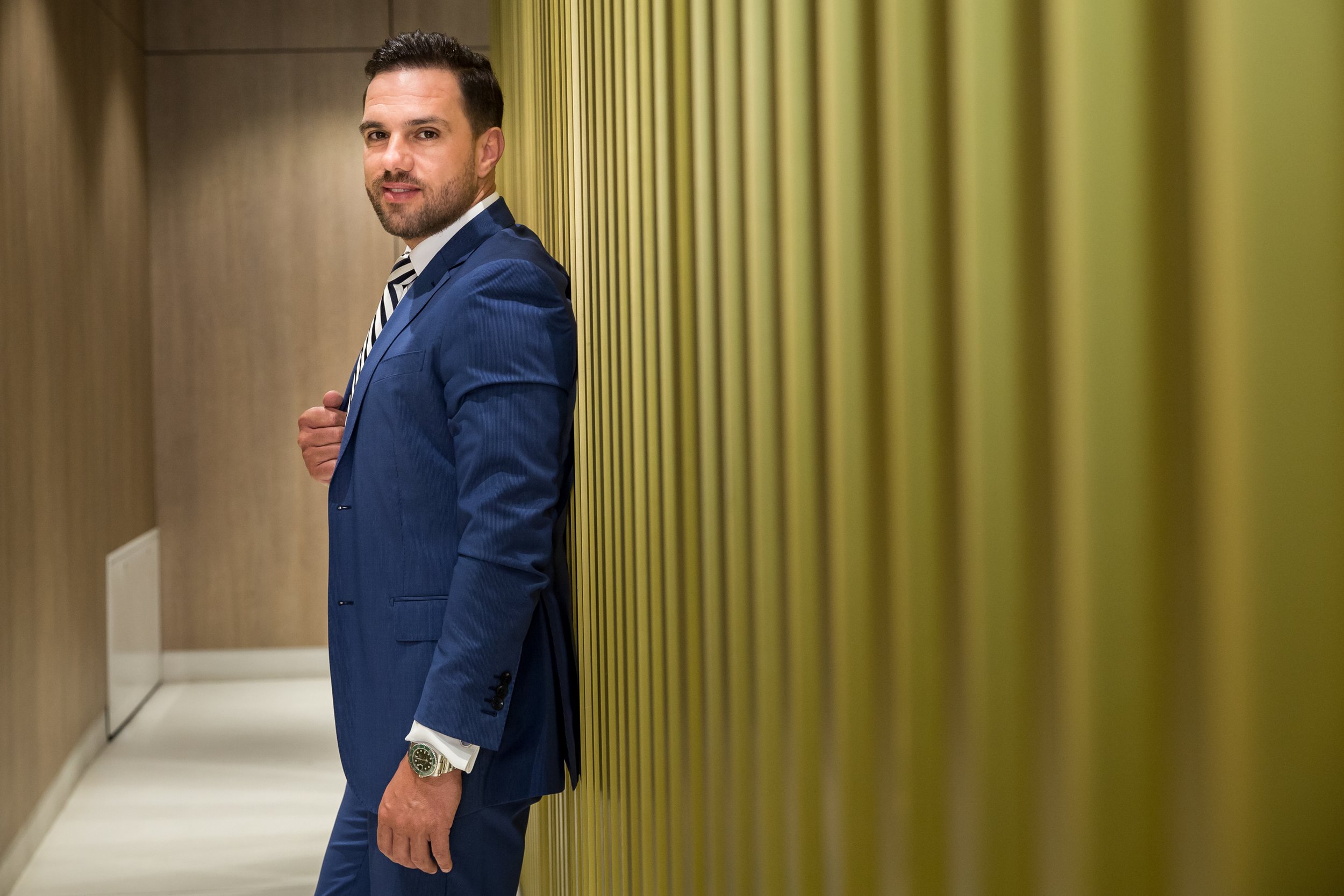 A man in a blue suit standing in a hallway near a yellow-green wall, looking at the camera.