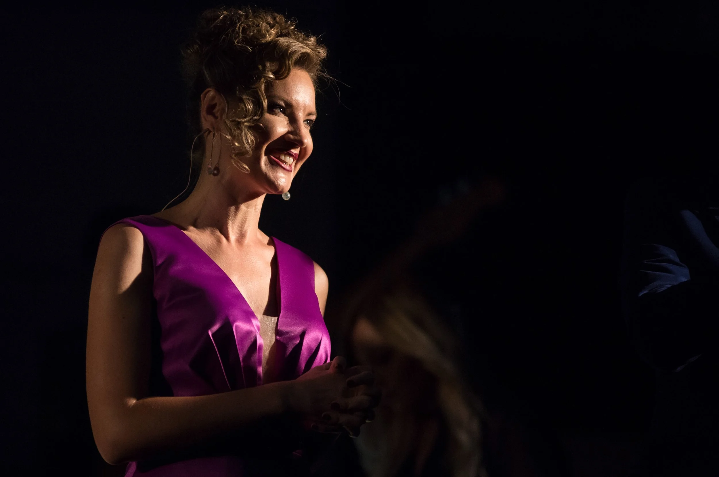 A woman wearing a purple dress, smiling and looking to her right, with curly hair and earrings, in a dark setting.