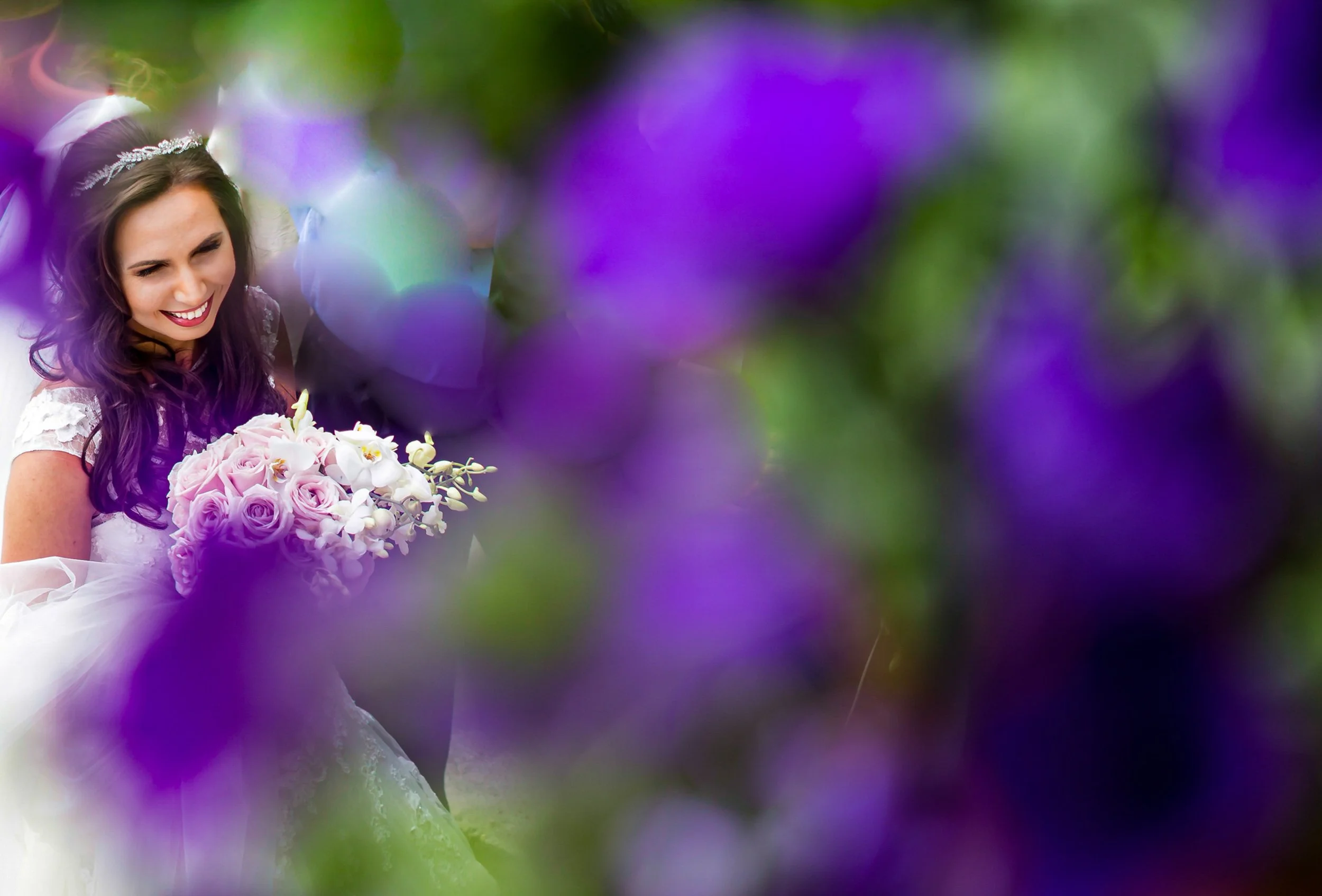 Smiling woman holding a bouquet of pink and white roses, decorated with orchids, at a wedding. She is wearing a tiara and a white wedding dress.