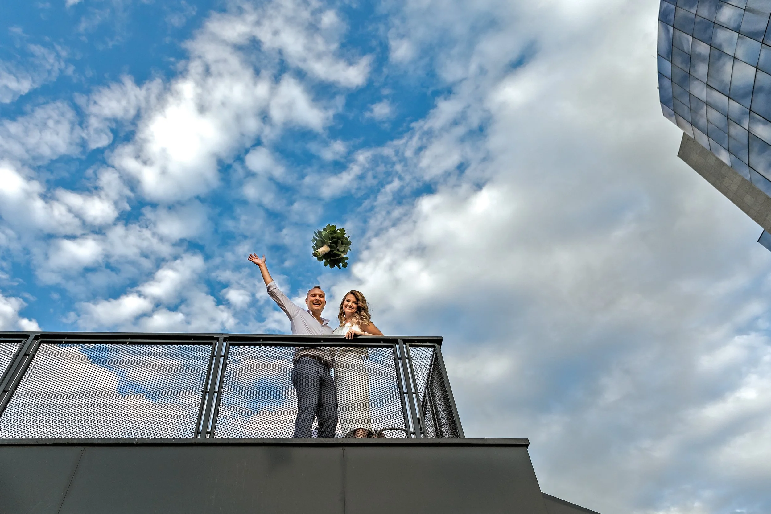 A couple celebrating on a rooftop, with the man holding a bouquet and throwing it into the air, against a background of a partly cloudy sky and modern glass building.