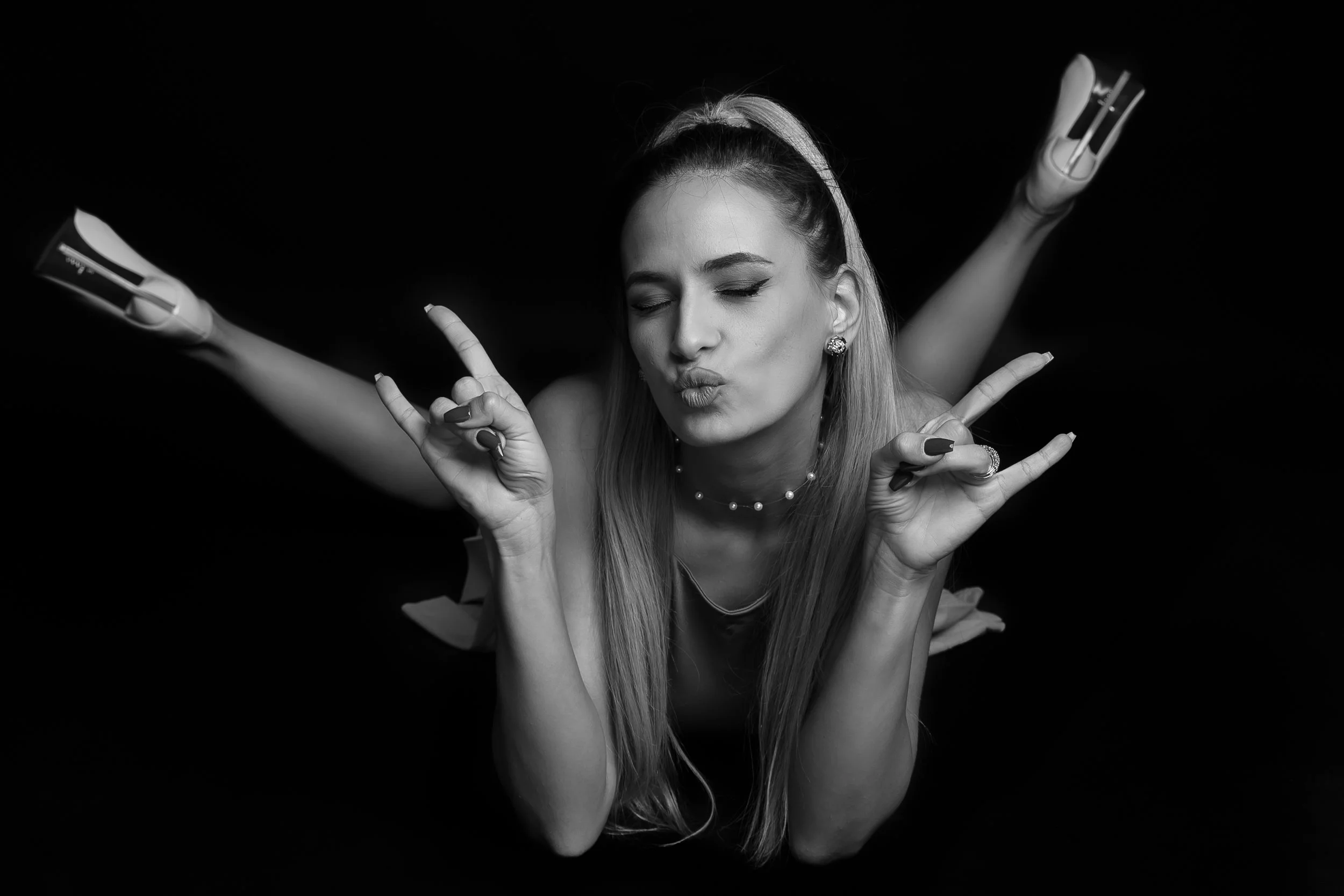 A young woman with long hair and a headband making a kissy face and peace signs with both hands. She is wearing earrings, a necklace, and a ring, with her eyes closed against a black background.
