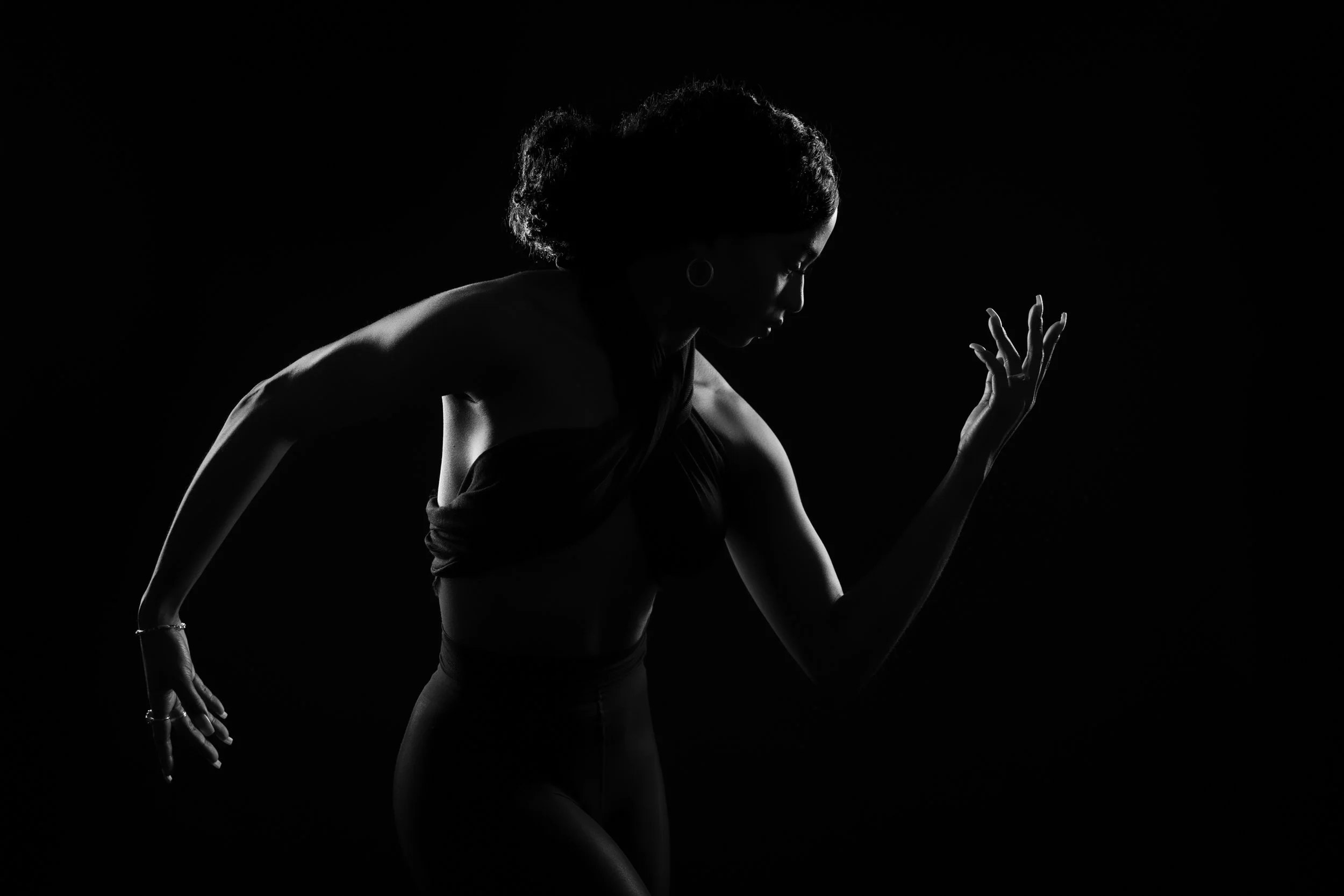A silhouette of a woman in a crouched pose with her hand raised, illuminated from the side creating a high contrast, black-and-white image.
