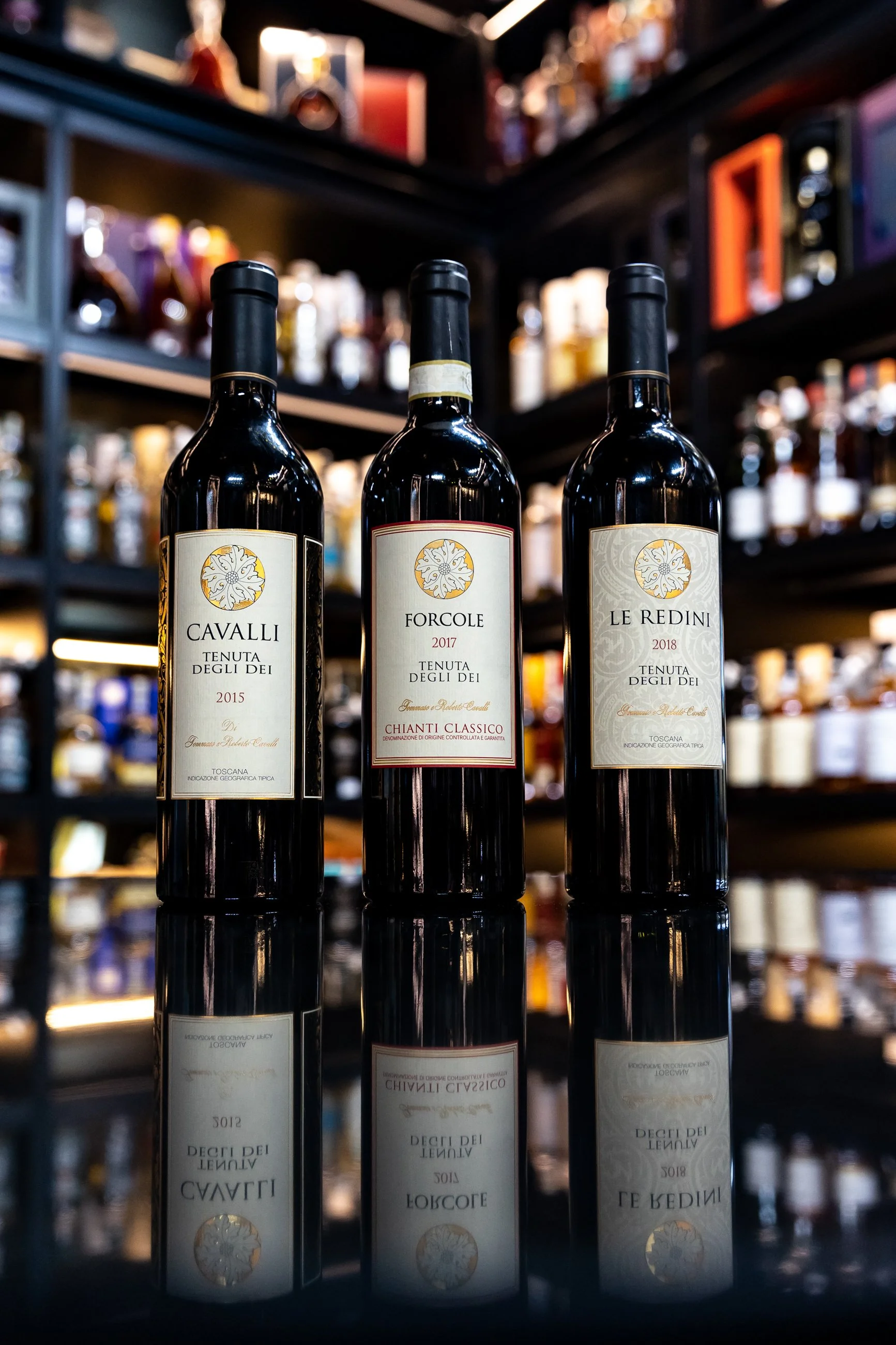 Three bottles of red wine with labels, on a reflective surface in a liquor store, with shelves of various liquor bottles and boxes in the background.
