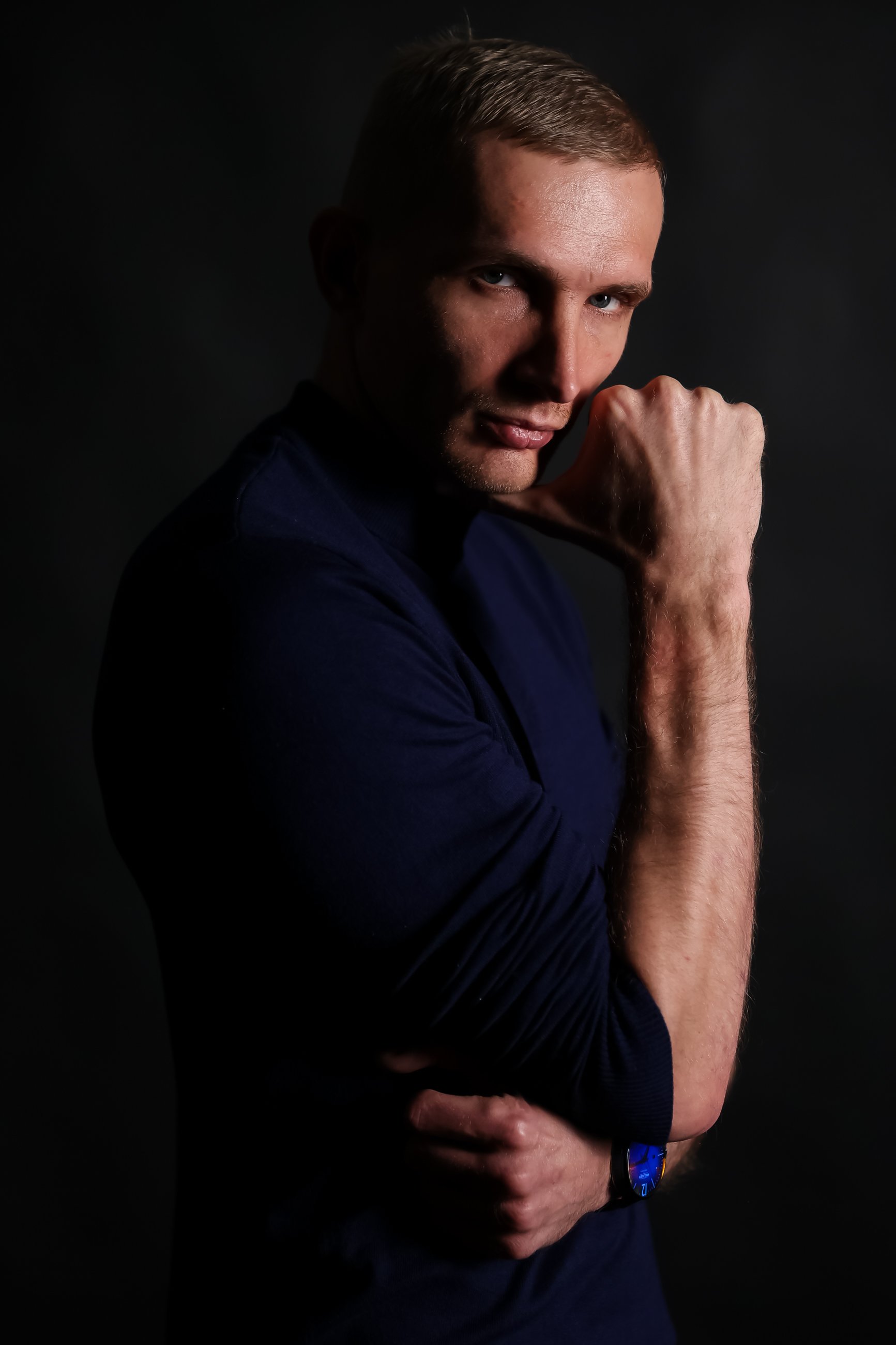 A young man with short brown hair, posing against a dark background, wearing a dark shirt, with his right hand clenched in a fist near his chin, and a serious expression on his face.