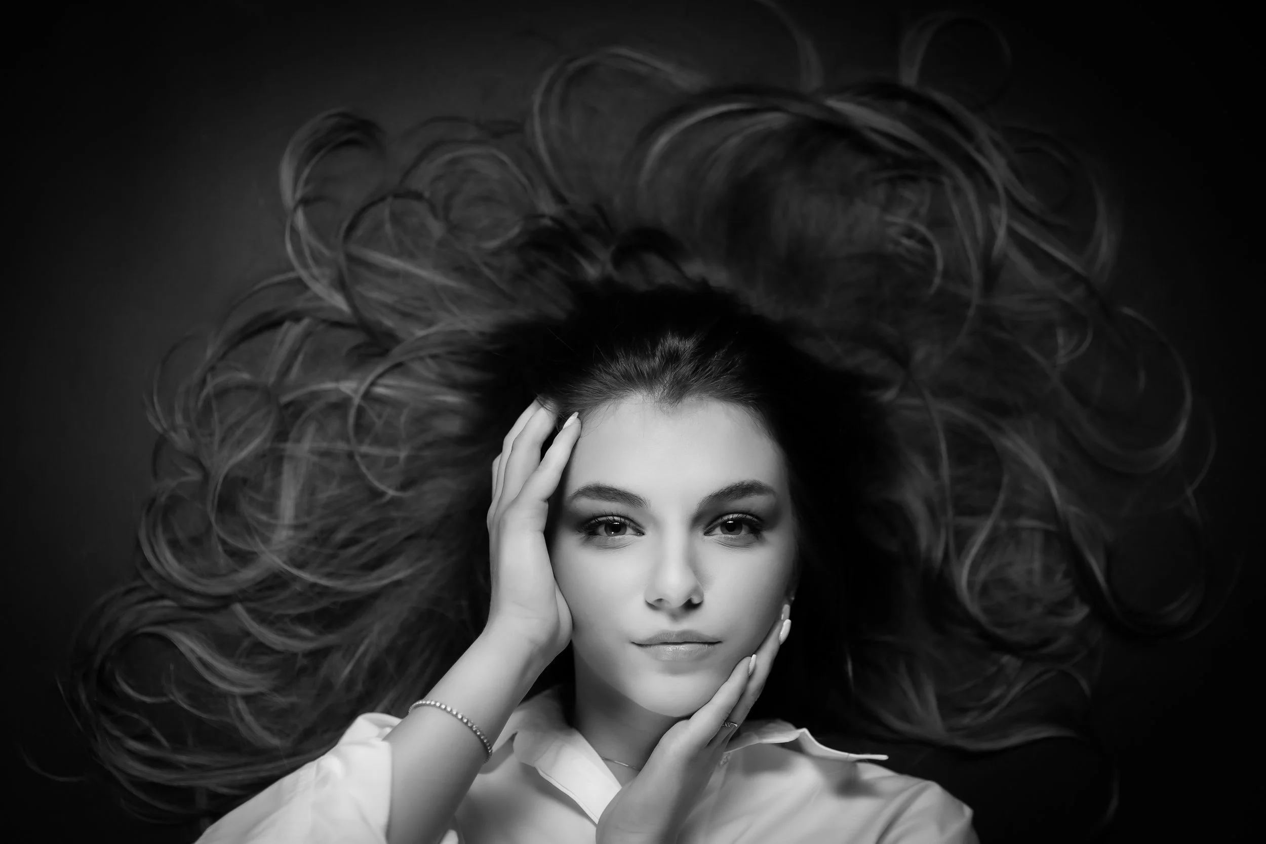 Black and white portrait of a woman with long, wavy hair spread out around her head, holding her face with both hands, wearing a white shirt and a bracelet.
