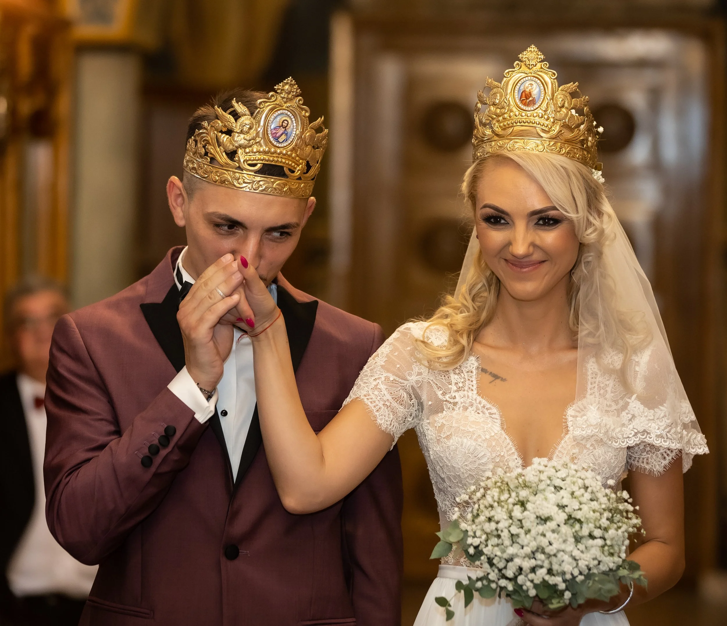 A bride and groom wearing ornate gold crowns during their wedding ceremony. The groom is dressed in a maroon tuxedo and the bride in a lace wedding dress, holding a bouquet of white flowers.