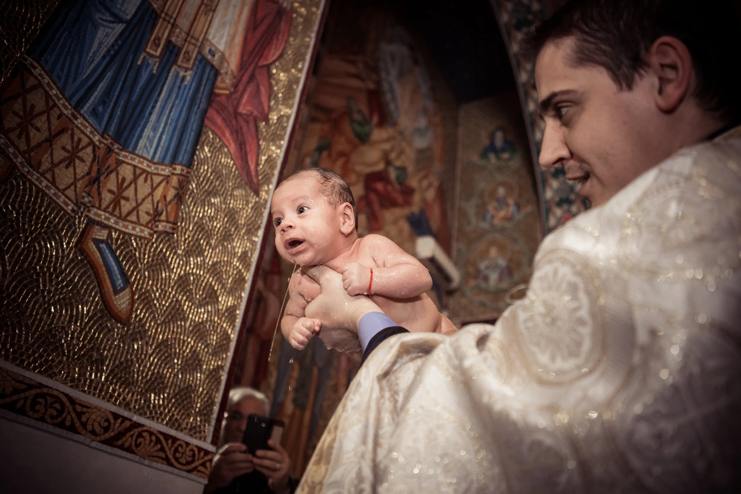 A priest or minister holding a baby during a religious baptism ceremony inside a church with ornate, colorful murals and gold accents.