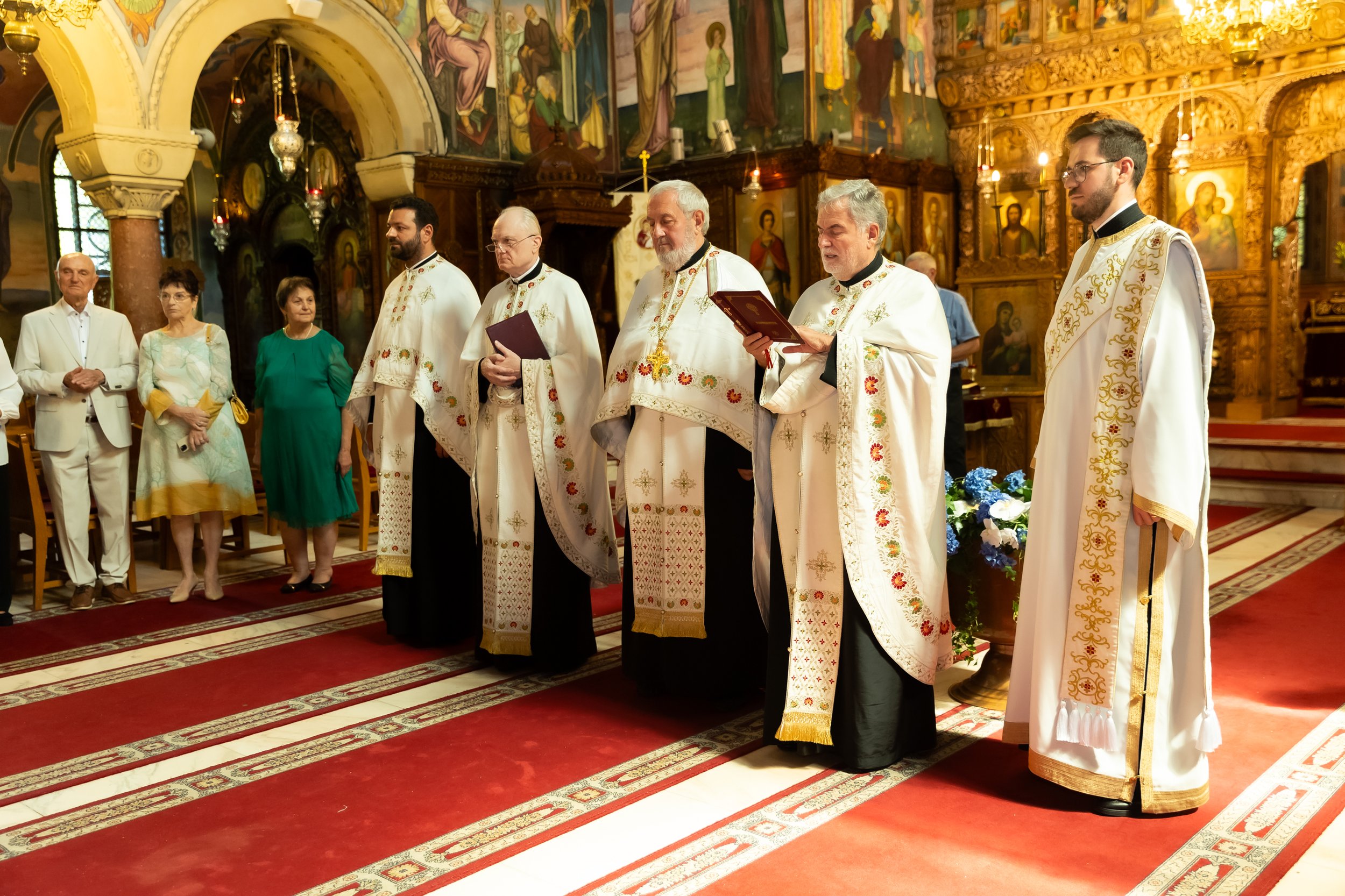 Priests in white embroidered vestments standing in a church interior with ornate wooden iconostasis and religious paintings, while a few people in the congregation stand nearby.