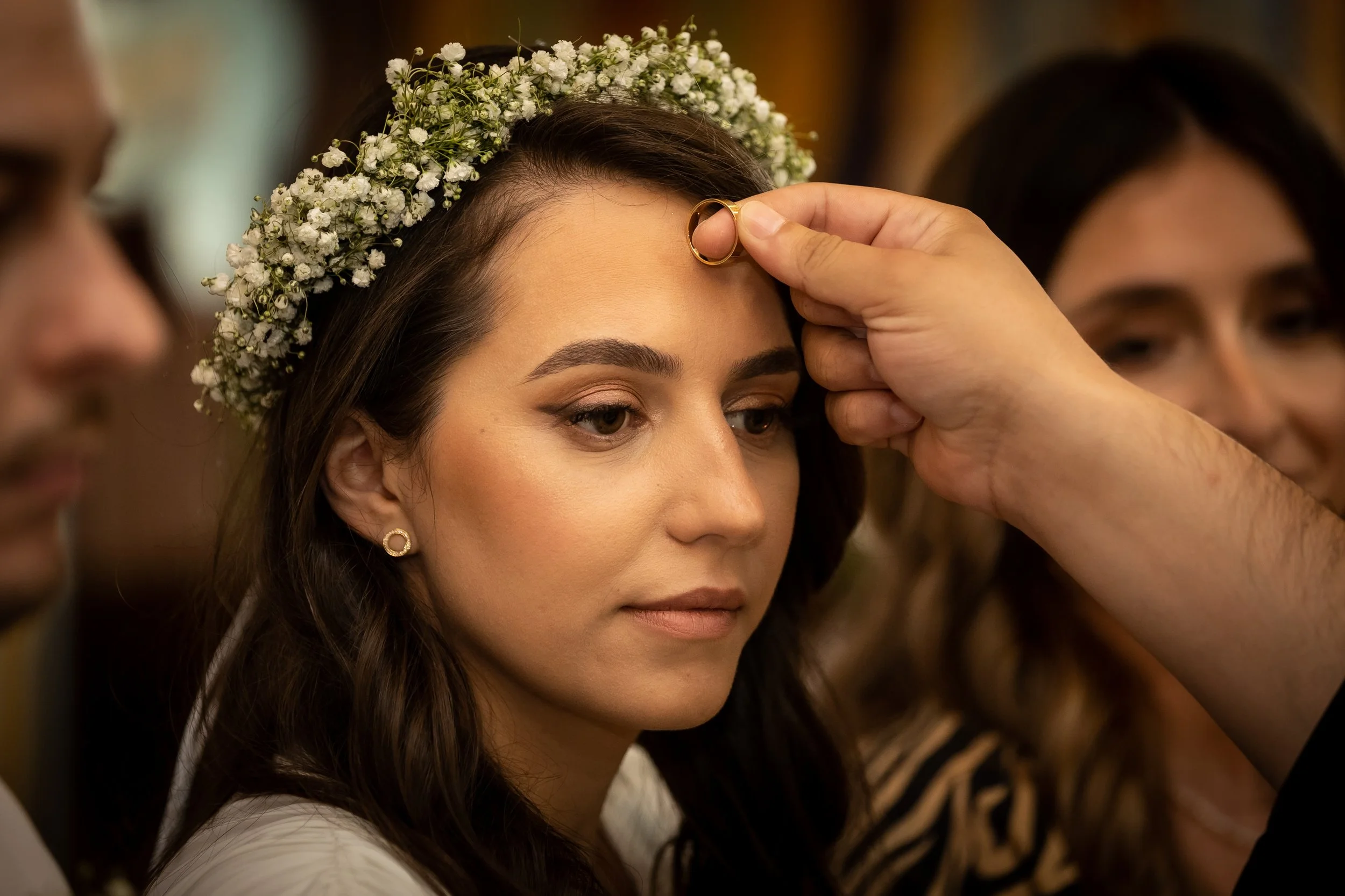 A woman with long brown hair and earrings has a flower crown placed on her head during a wedding ceremony. Someone is putting a gold ring on her finger.