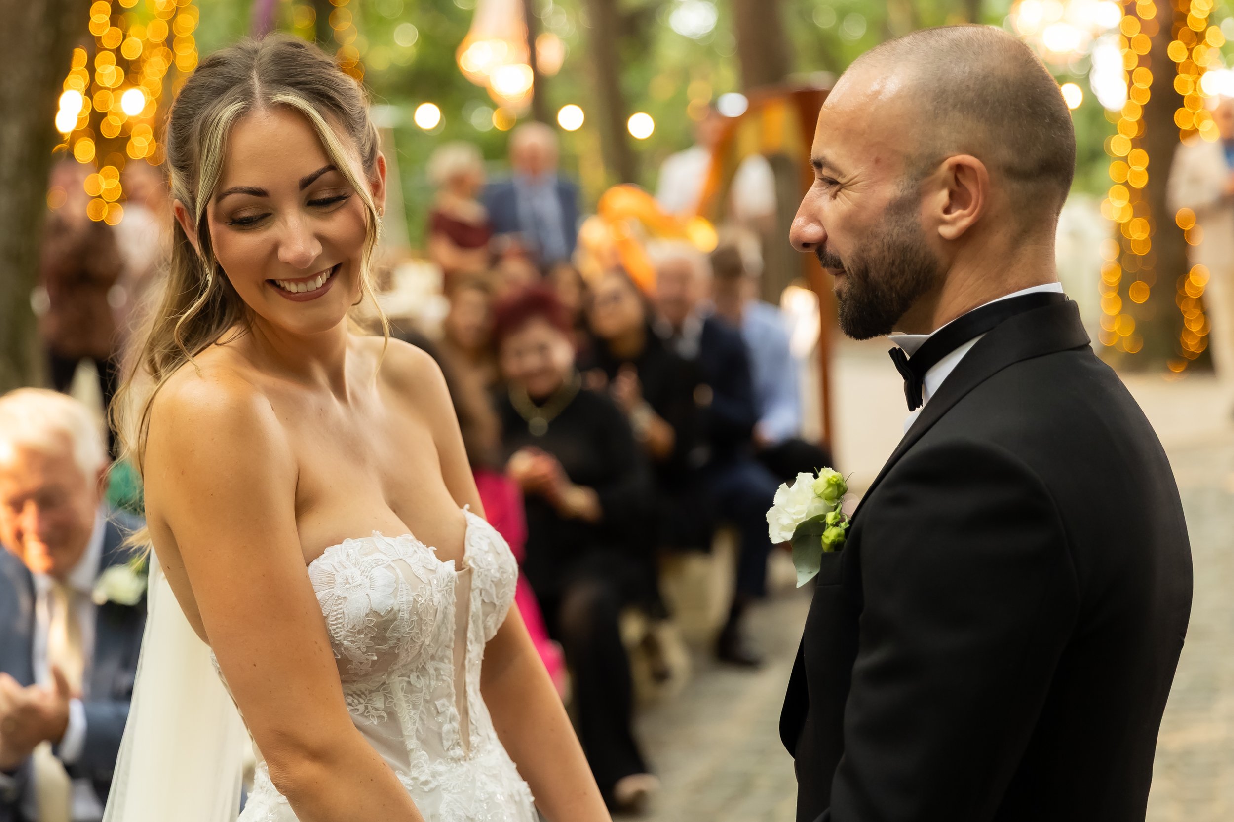 A bride and groom during their outdoor wedding ceremony, exchanging vows, with guests in the background and decorative string lights hanging in the trees.