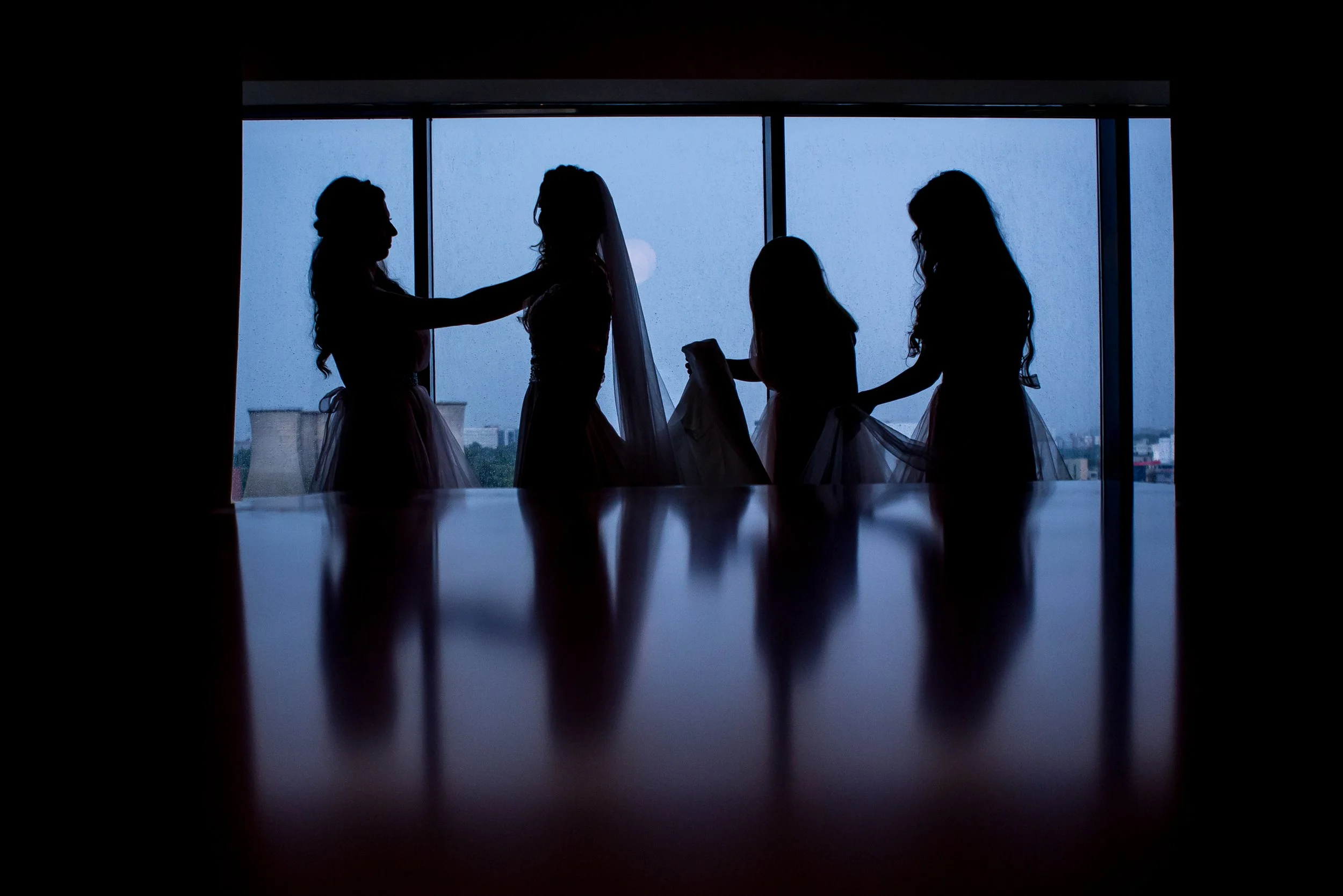 Silhouettes of four women dressed in formal attire, standing near a large window with a foggy cityscape in the background, possibly during a wedding preparation.