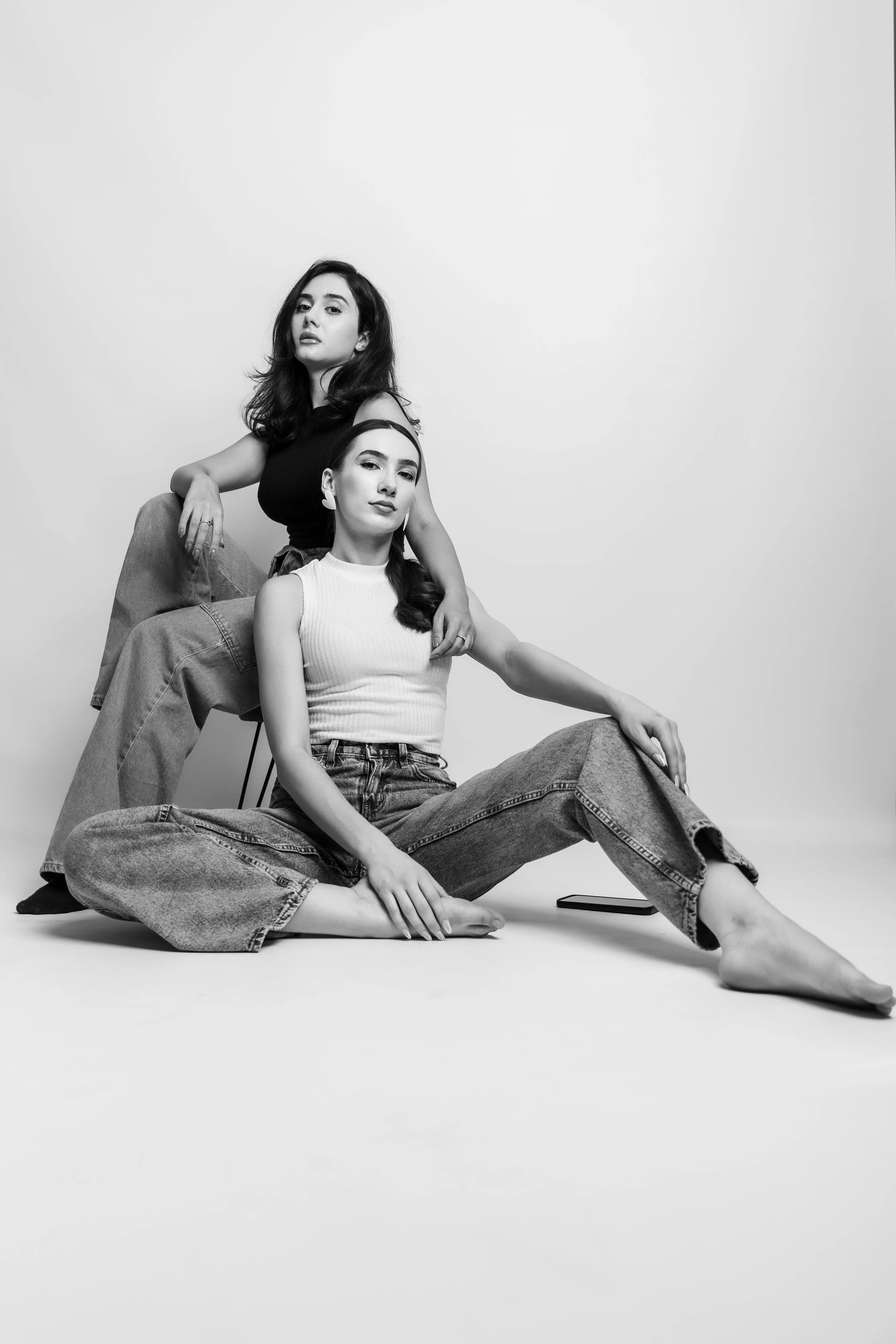 Two women posing together in black and white. One woman is sitting on the floor with her legs crossed, wearing a sleeveless top and jeans, with long dark hair. The other woman is seated on a chair with one leg on the floor and the other draped over t