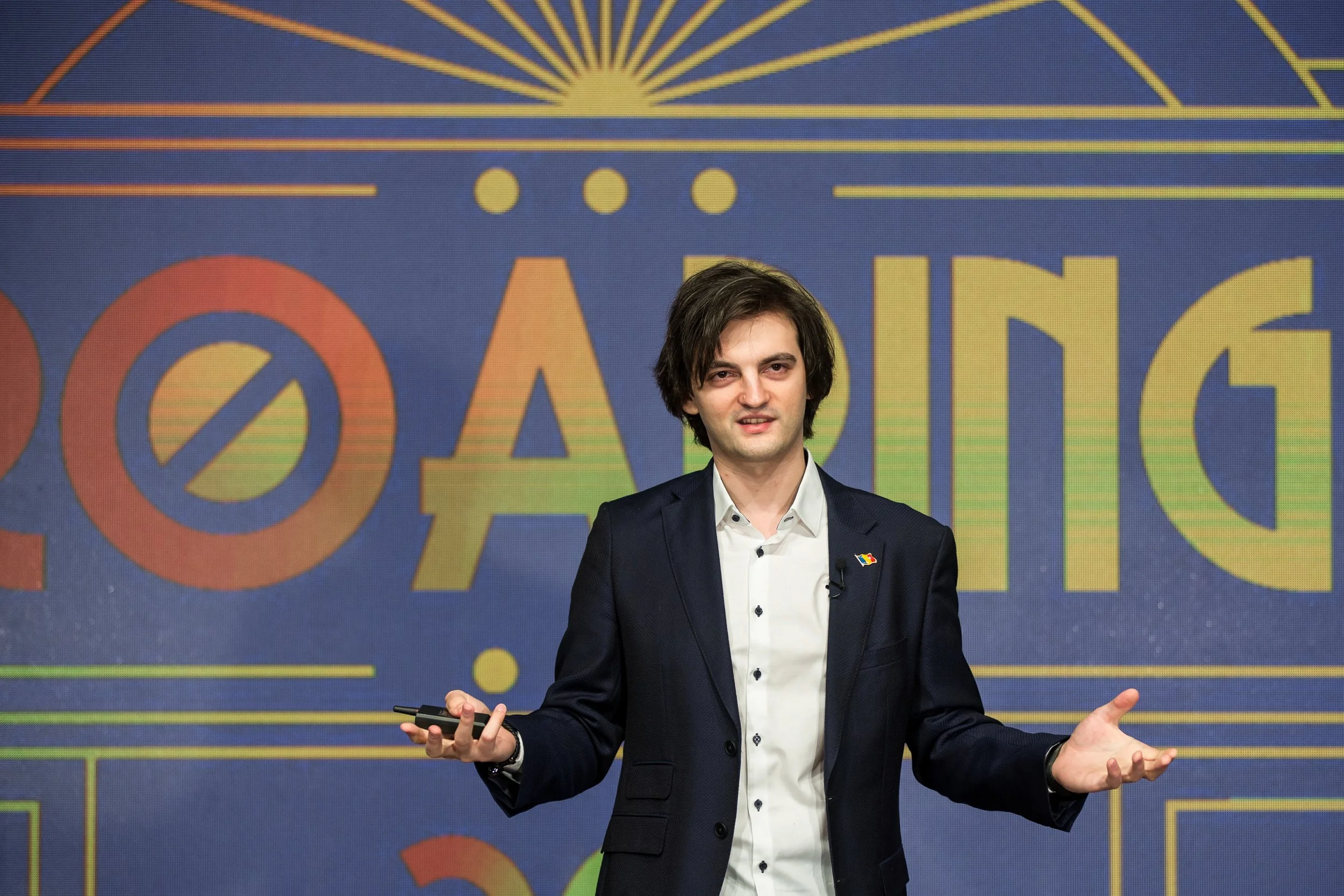A man in a dark suit and white shirt, gesturing with his right hand, standing in front of a colorful background with the word 'Voting' and the symbol for no or prohibition.