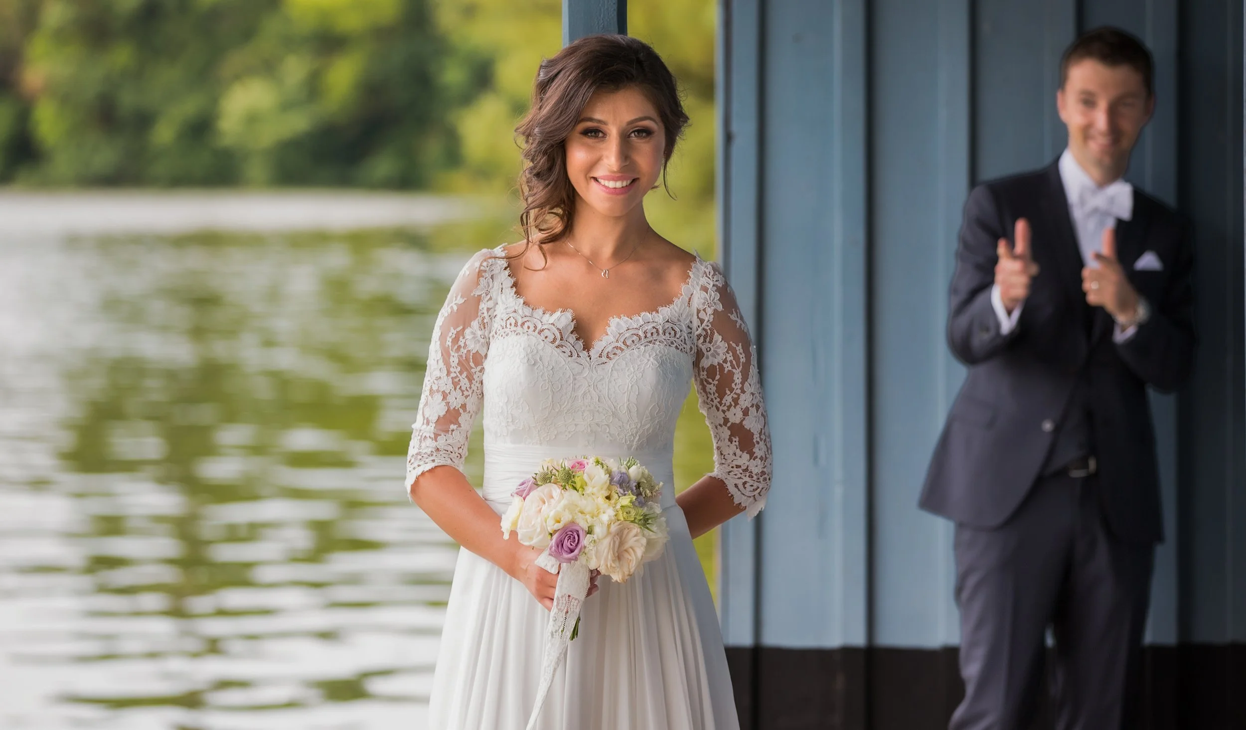 A bride in a white lace wedding dress holding a bouquet of flowers, standing outdoors near a body of water with a groom in a dark suit visible in the background giving a thumbs-up.