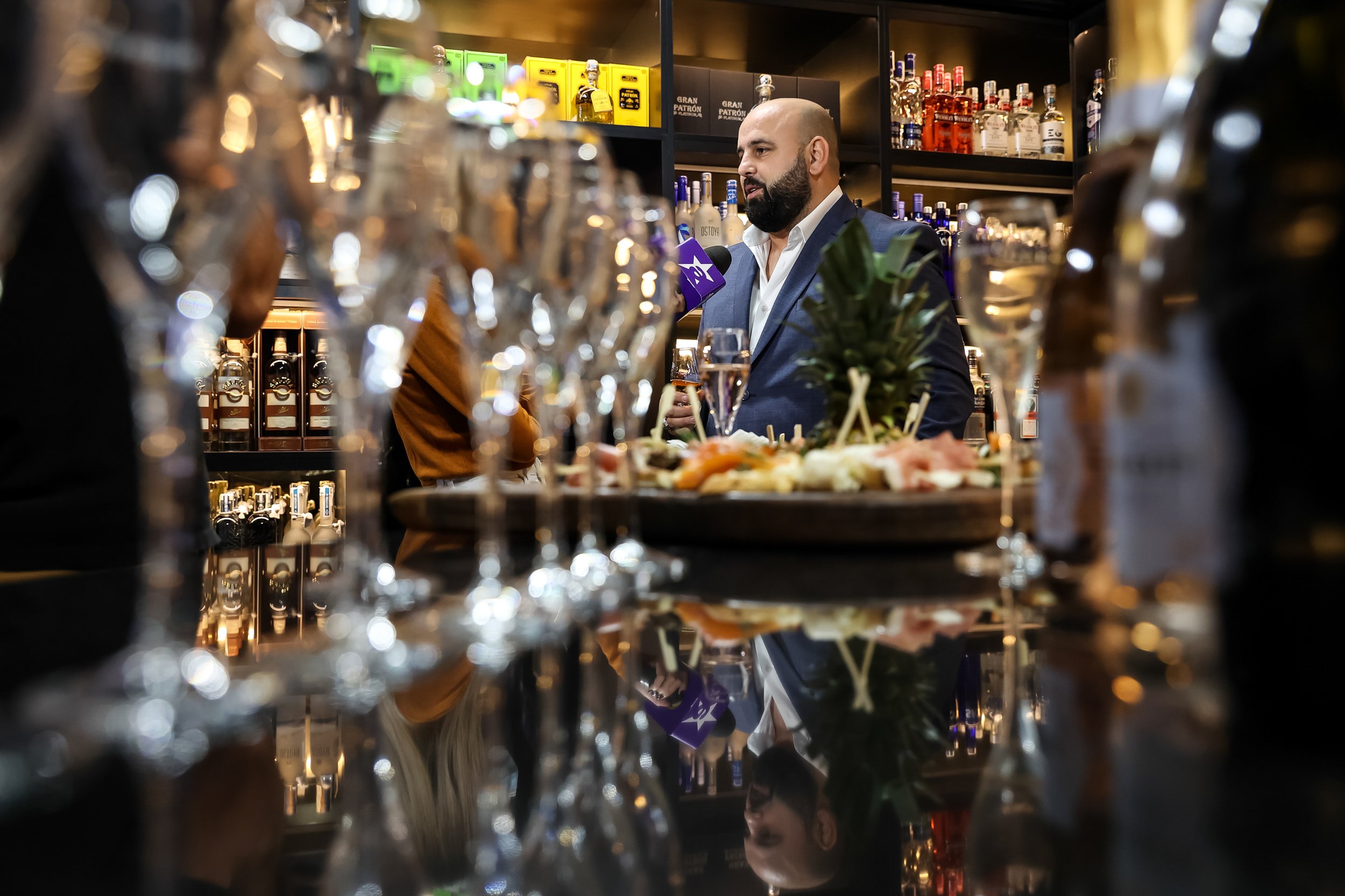 A man in a suit and white shirt, with a beard, is behind a bar, holding a drink. The bar is decorated with various bottles of alcohol, glasses, and a small potted plant. The background shows shelves stocked with bottles of liquor and boxes, with a fo