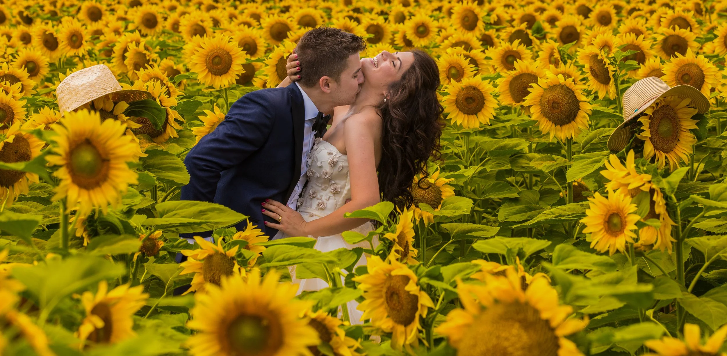 A couple in wedding attire kissing in a sunflower field with sun hats on some sunflowers in the background.