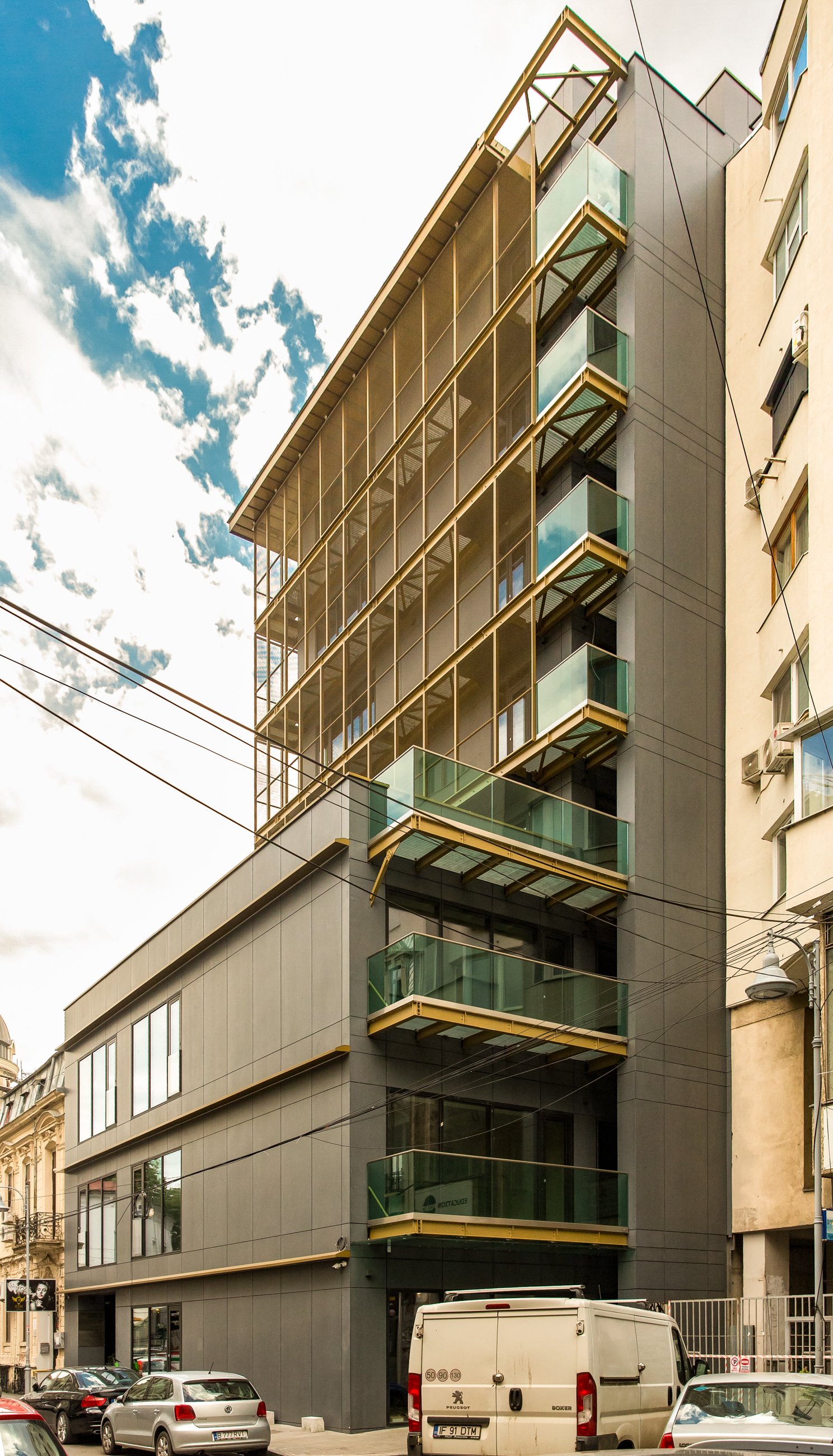 Modern multi-story building with glass balconies, situated in an urban area with parked cars in front.