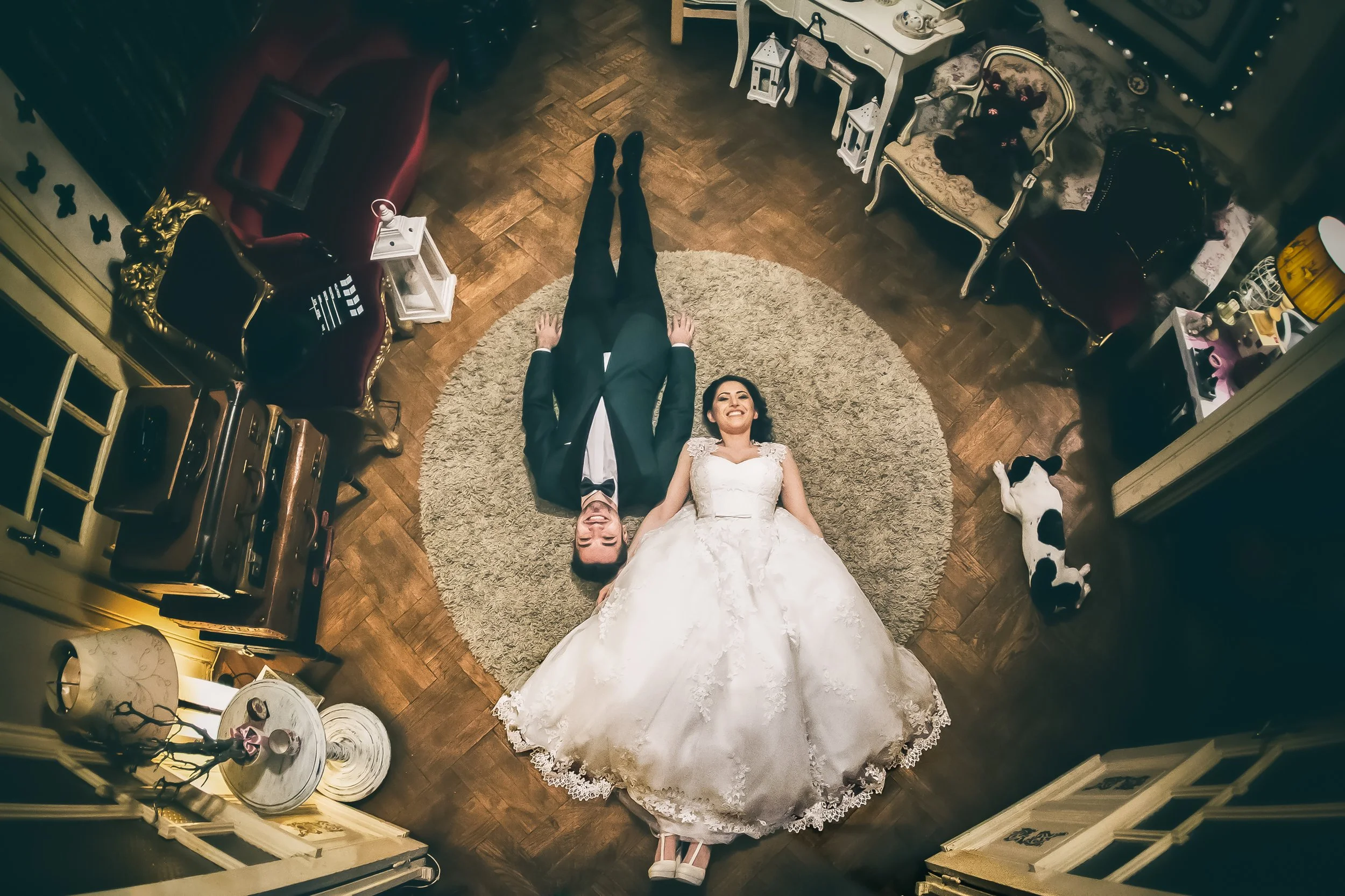 A bride and groom lying on the floor of a vintage styled room, smiling and holding hands, with the bride in a white wedding dress and the groom in a dark suit. The room has wooden flooring, ornate chairs, and vintage decoration.