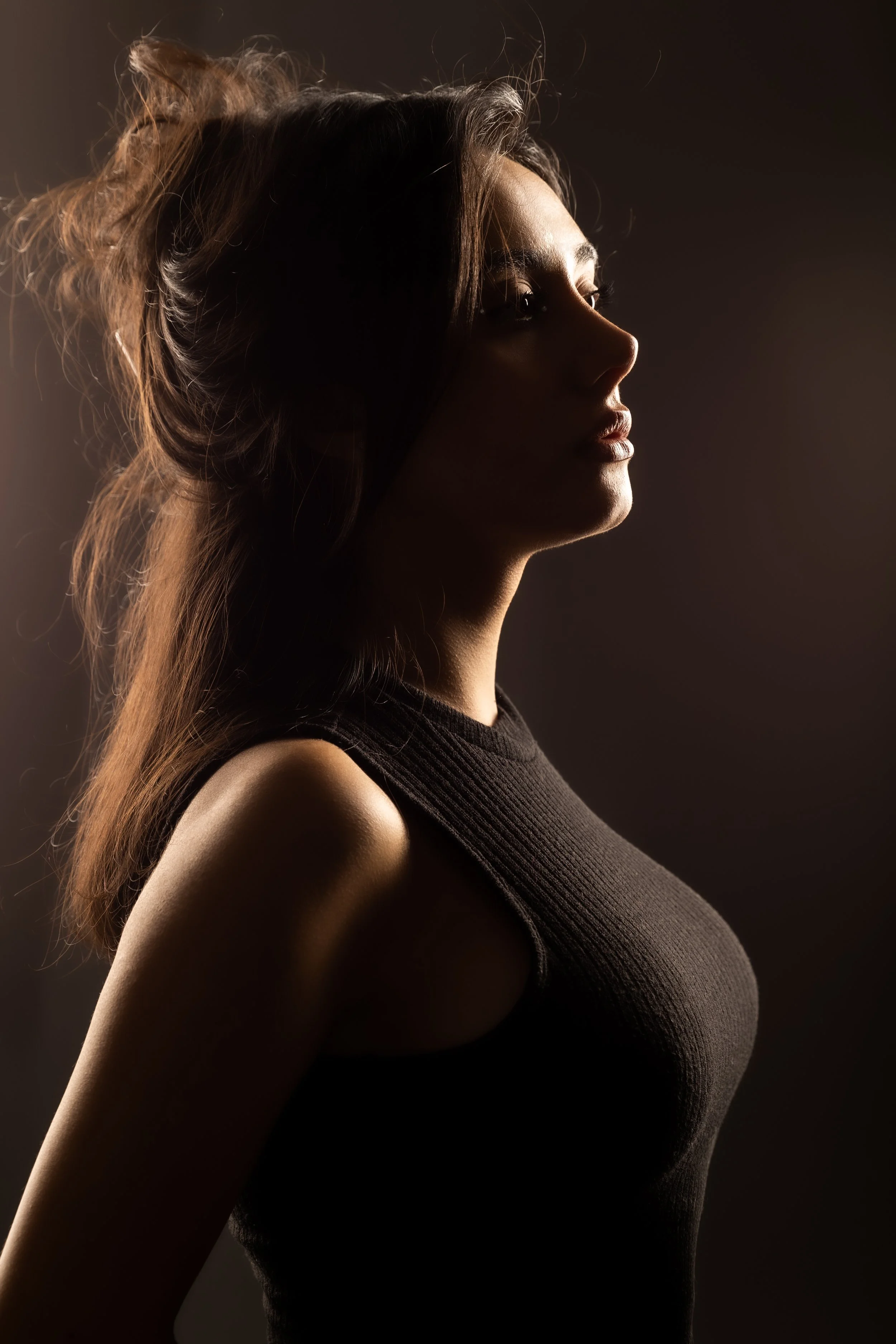 Profile of a woman with dark hair in a loose bun, wearing a black sleeveless top, with soft lighting highlighting her face and shoulder against a dark background.