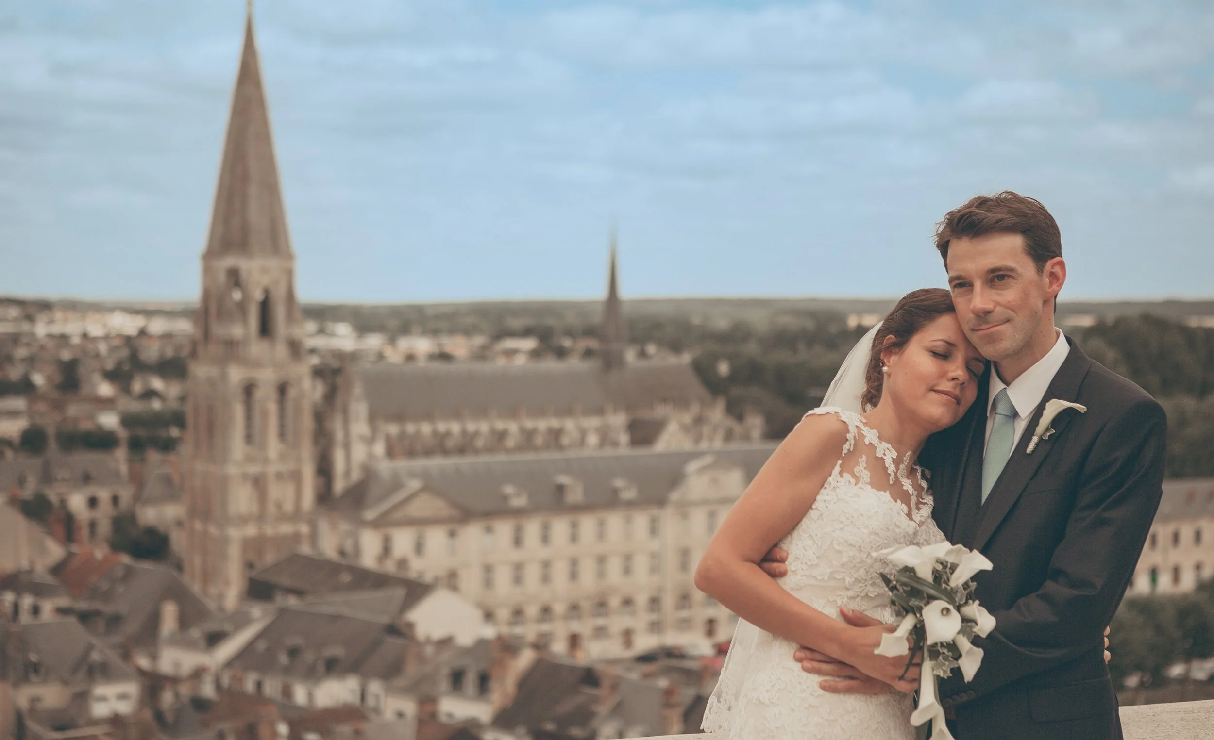 A bride and groom embrace on a rooftop with a European cityscape and church spires in the background.
