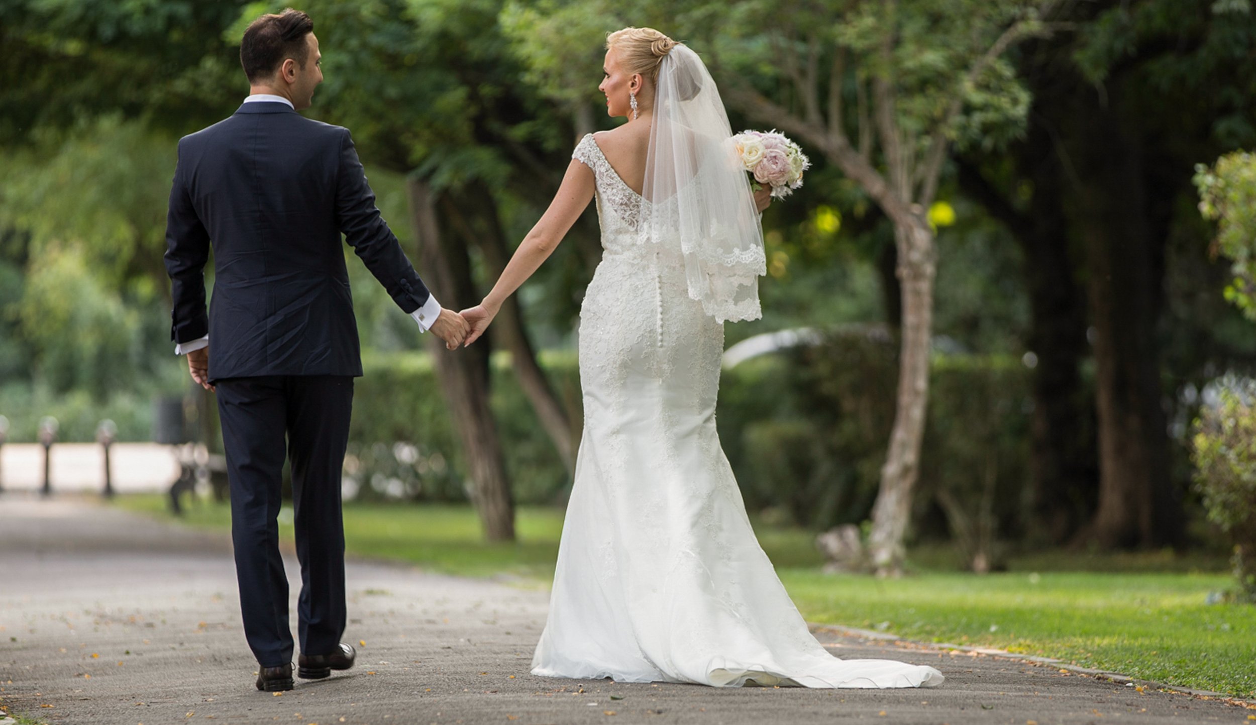 A bride and groom holding hands while walking outdoors in a park, surrounded by trees, during their wedding.