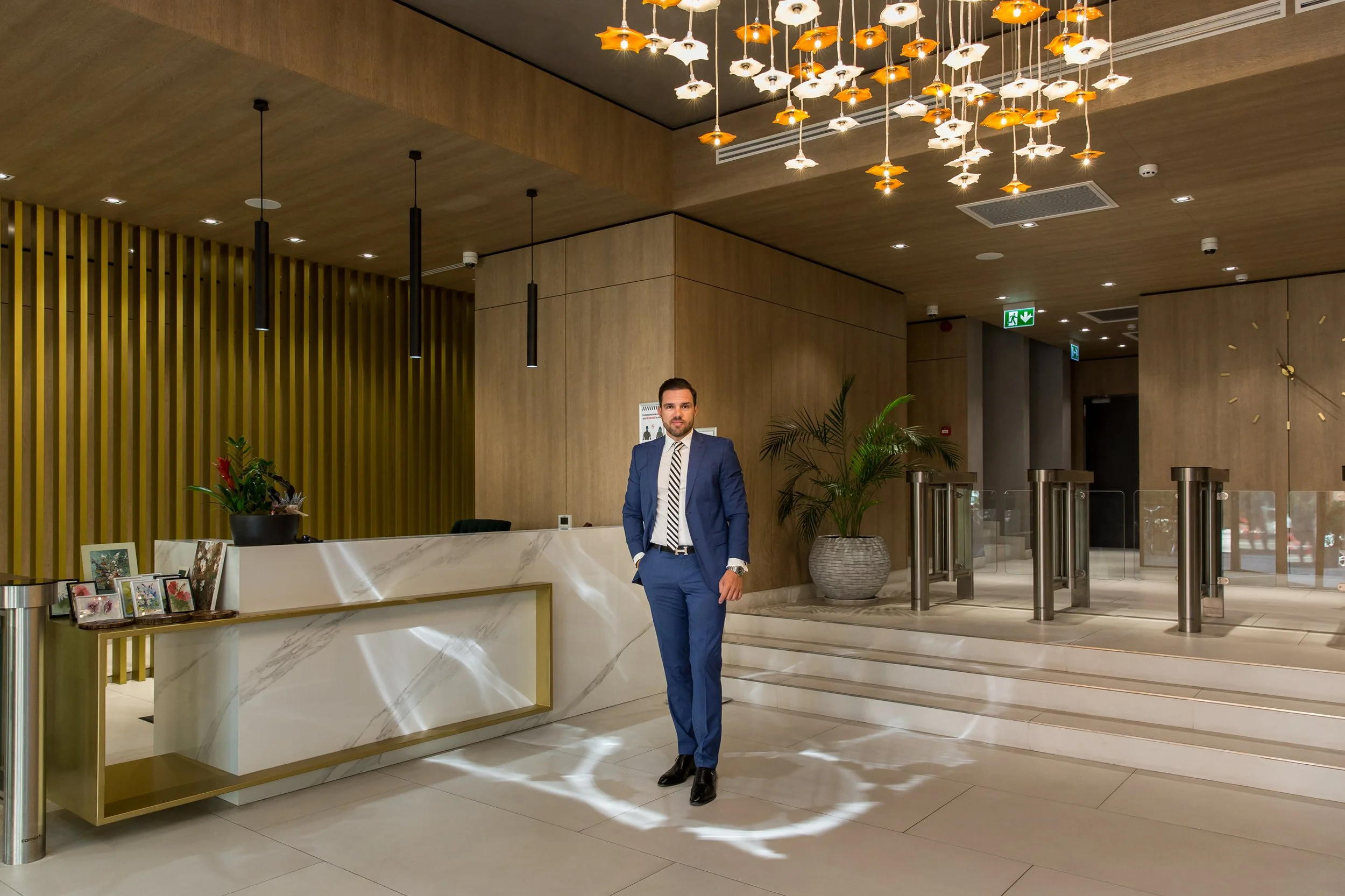 A man in a blue suit and striped tie stands in a hotel lobby, next to a white marble reception desk decorated with small framed pictures and a potted plant. The lobby features wood-paneled walls, modern pendant lighting with fish-shaped covers, and l