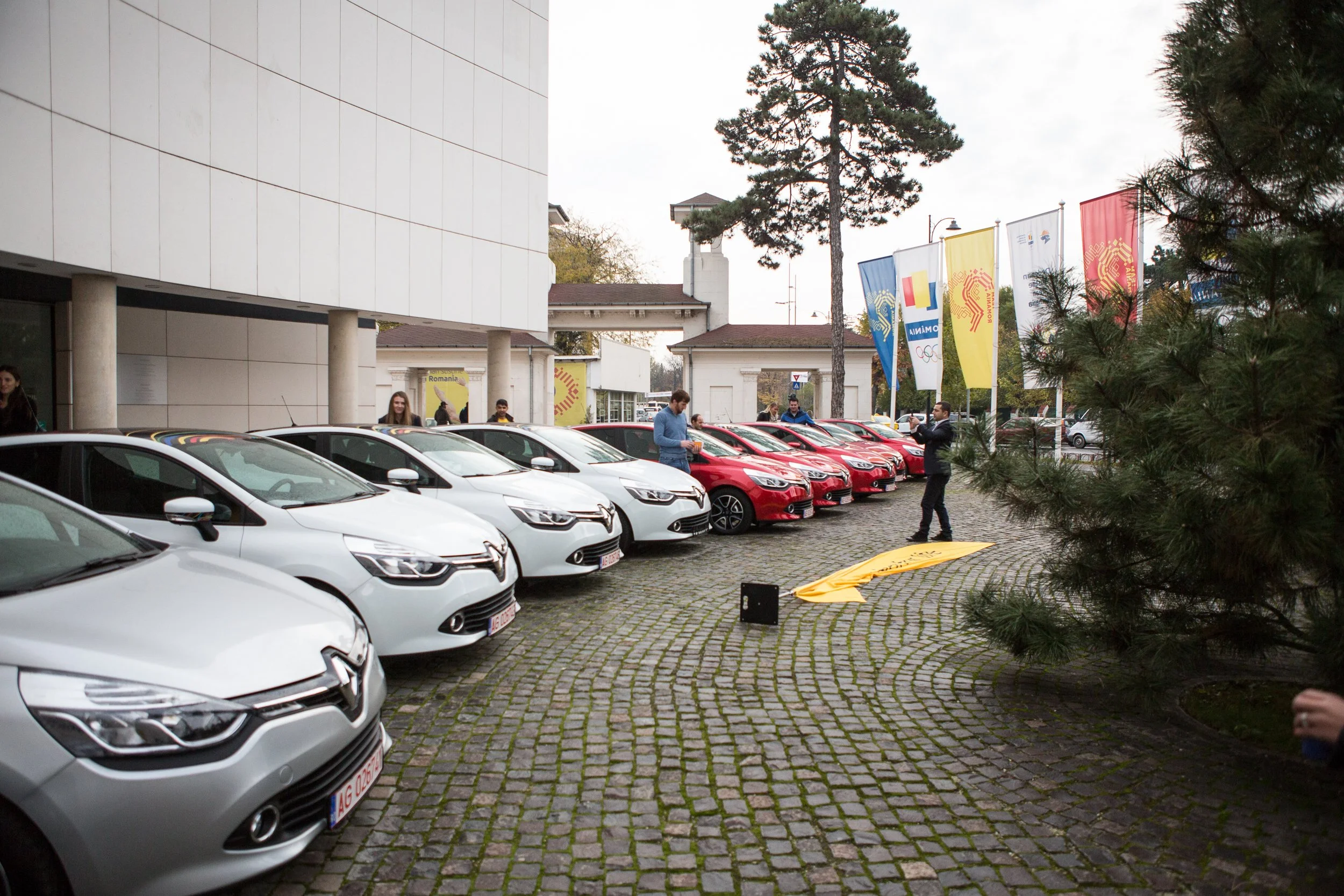 A parking lot with multiple white and red cars, people standing and talking near the cars, flags with Olympic and Romanian symbols, and a large tree in the background outside a modern building.