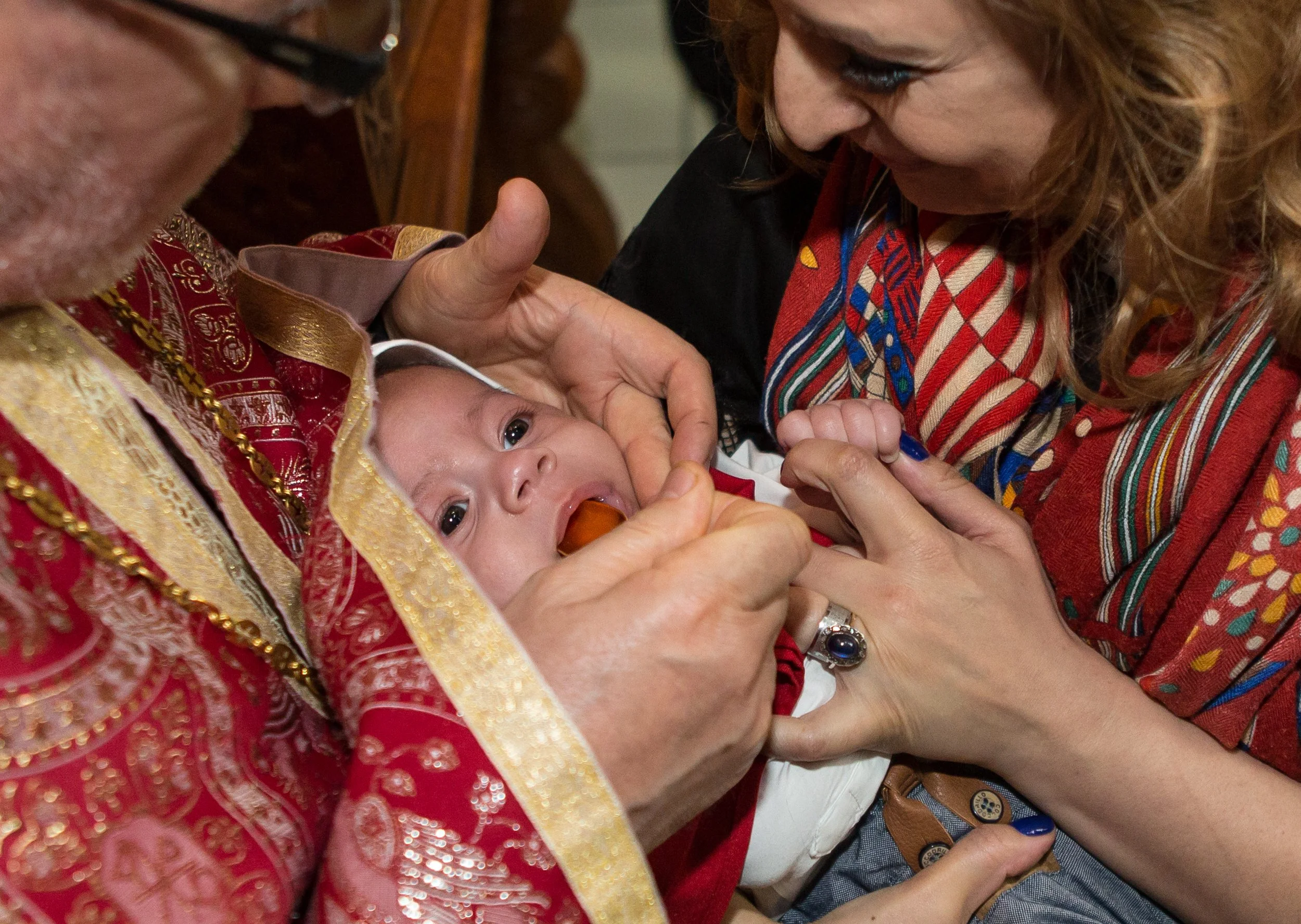 Close-up of a baby receiving a religious blessing or baptism from woman dressed in colorful traditional clothing, with adult beside them, part of a person wearing glasses visible on the left.