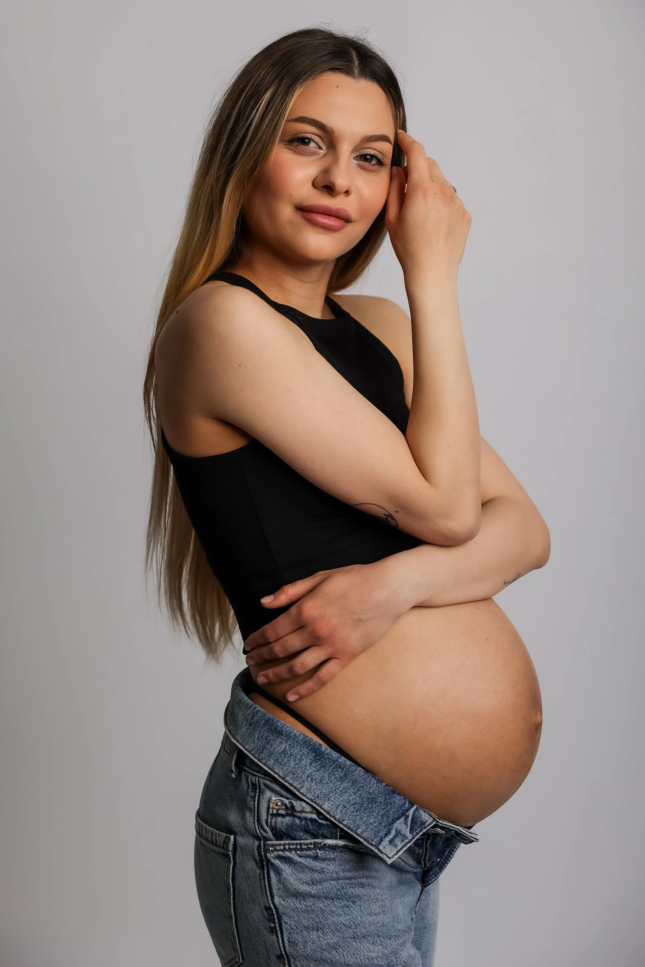 A pregnant woman with long blonde hair, wearing a black tank top and blue jeans, standing against a plain gray background. She is smiling slightly, with her left hand resting on her face and her right arm across her stomach.