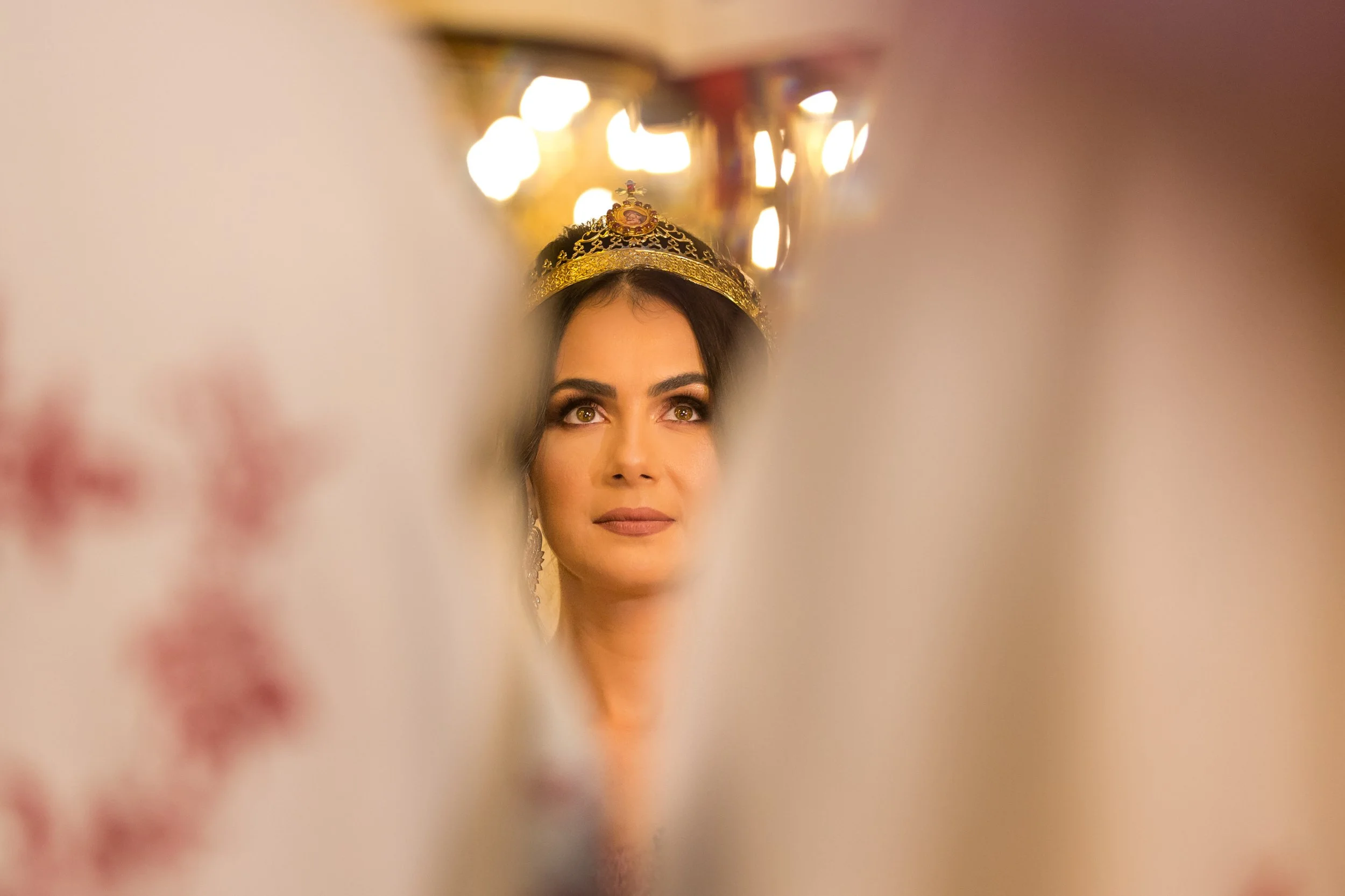 A woman with dark hair and makeup wearing a gold ceremonial crown, seen through a blurry foreground with warm lighting.