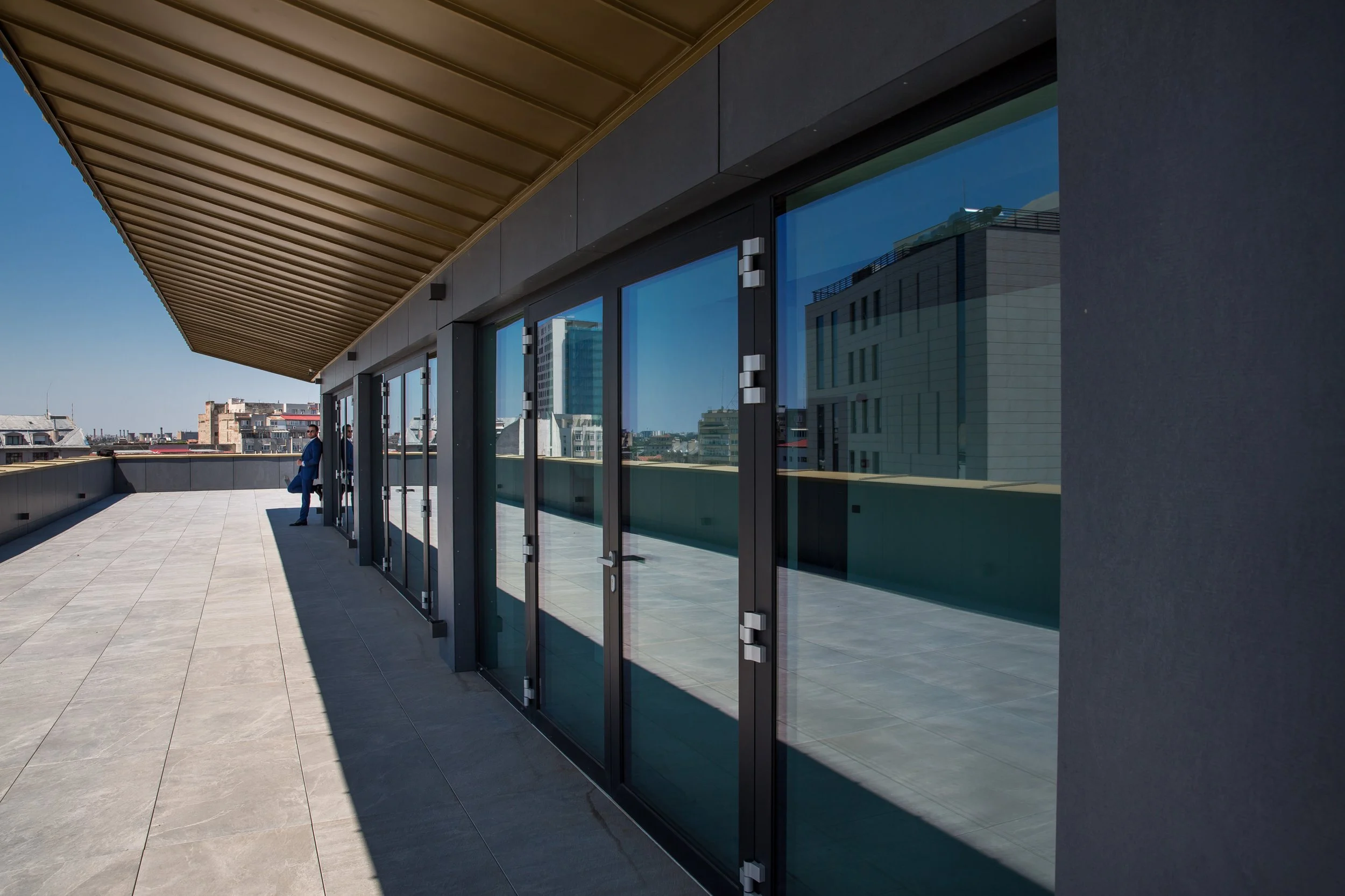A modern rooftop terrace with large glass doors, an overhanging roof with a tan underside, and city buildings in the background. Two people are standing near the doors.