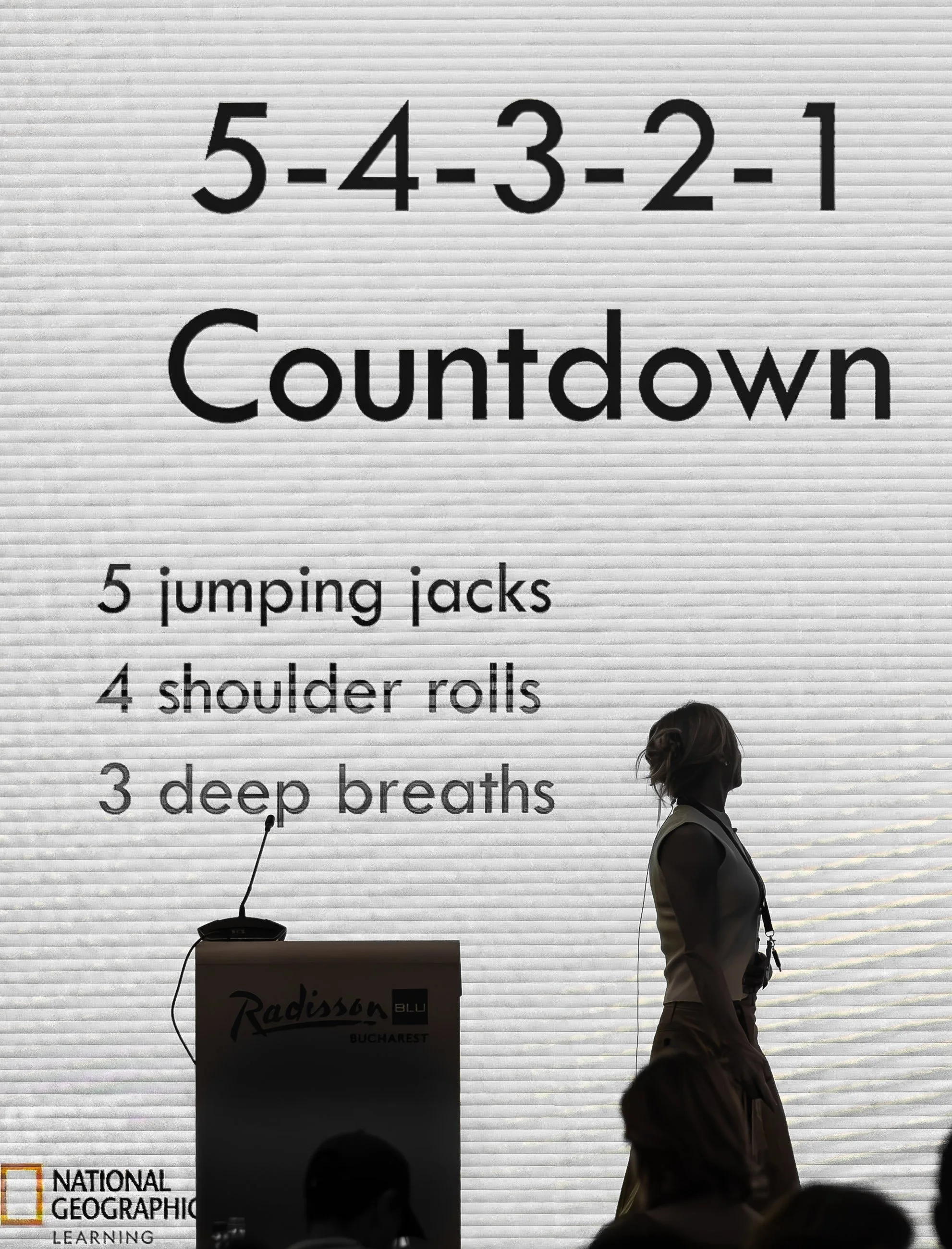 A woman stands in front of a large display screen with a countdown that shows the numbers 5, 4, 3, 2, 1 in sequence. The screen displays instructions for a workout, including jumping jacks, shoulder rolls, and deep breaths. The image is taken at the 