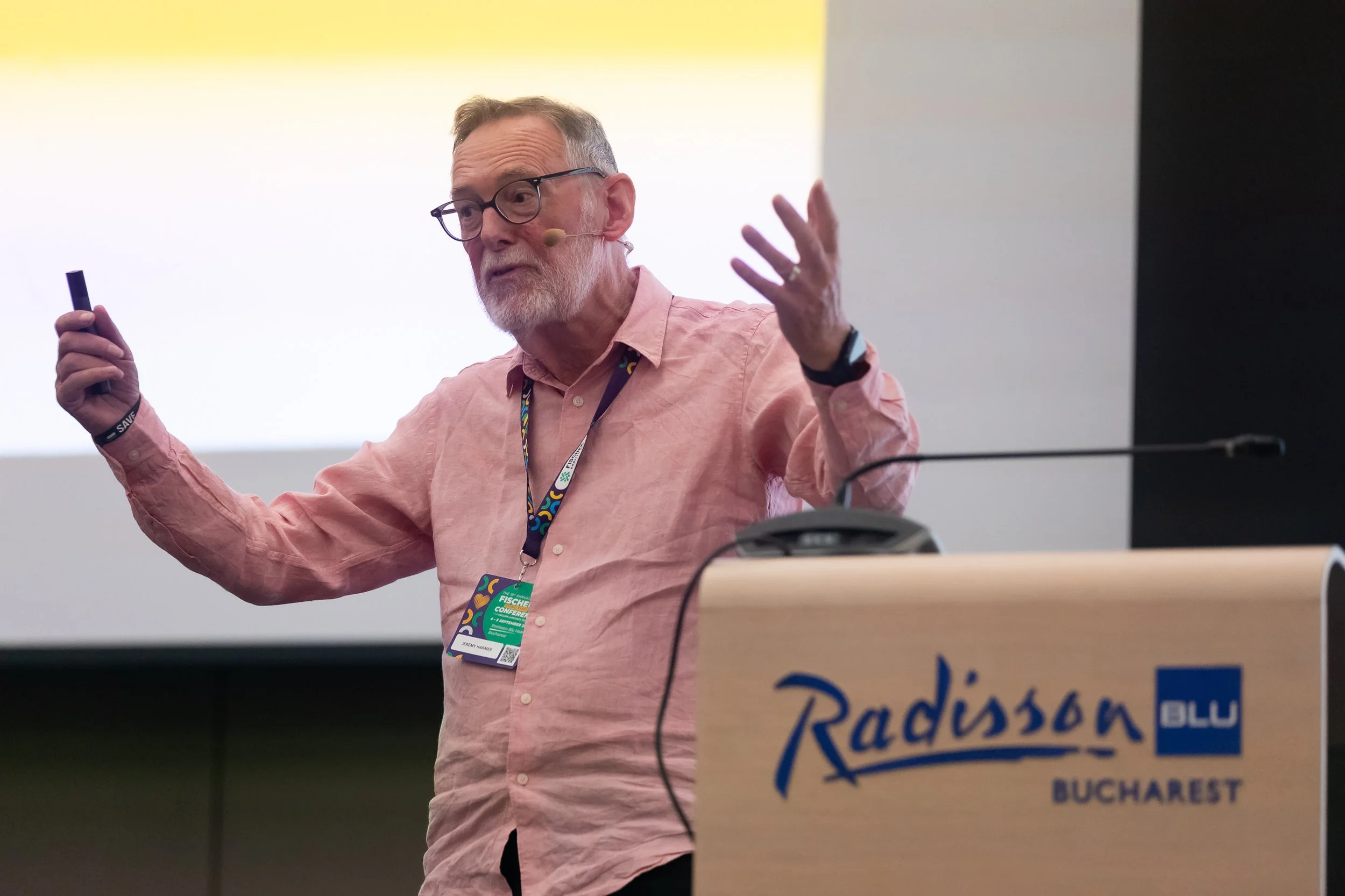 A man with glasses and a beard, wearing a pink shirt and a conference badge, is gesturing with his hands while speaking at a podium with a sign reading "Radisson Bucharest."