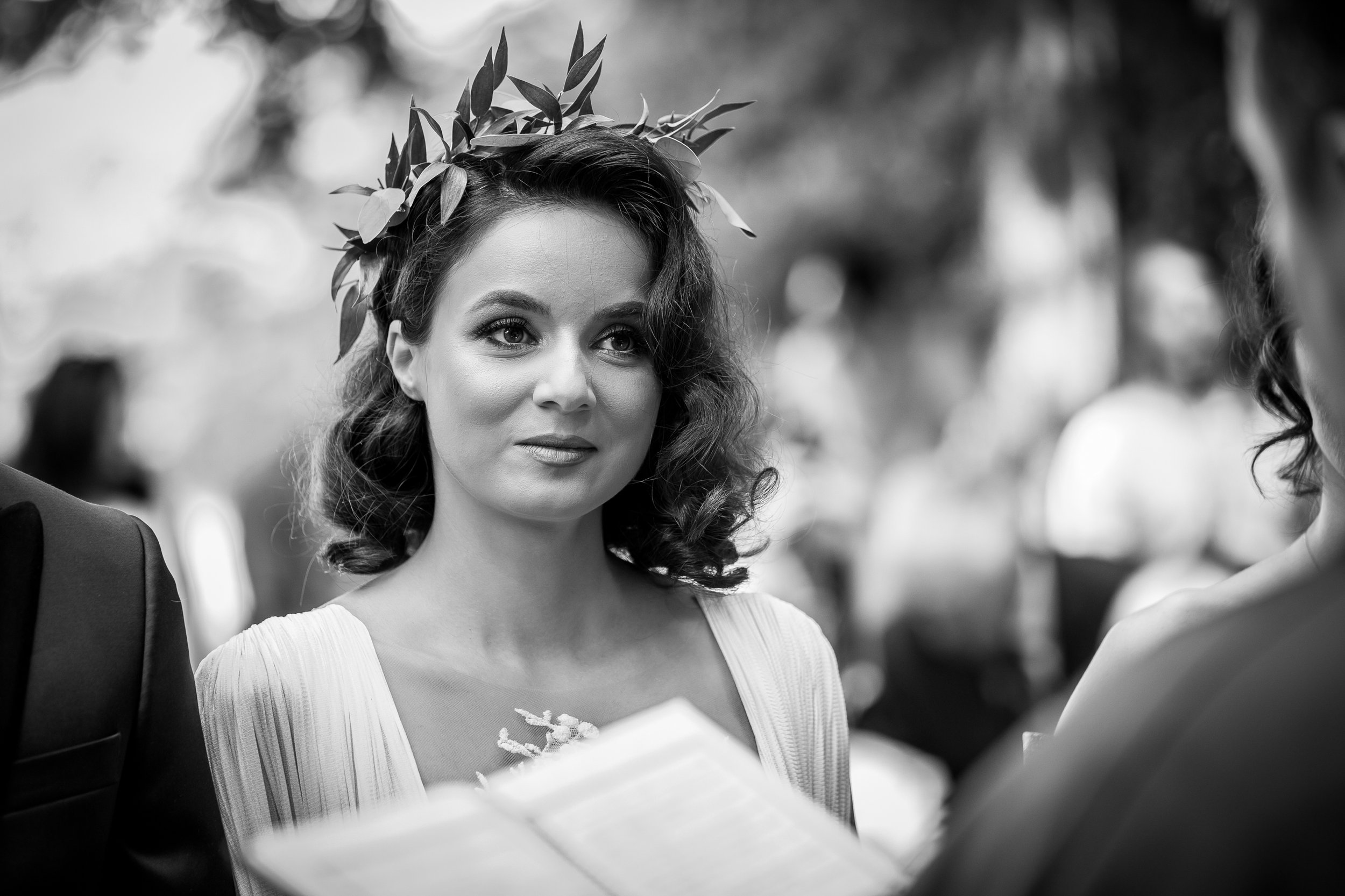 A woman with curly hair wearing a crown of leaves, dressed in a white gown, at an outdoor event.