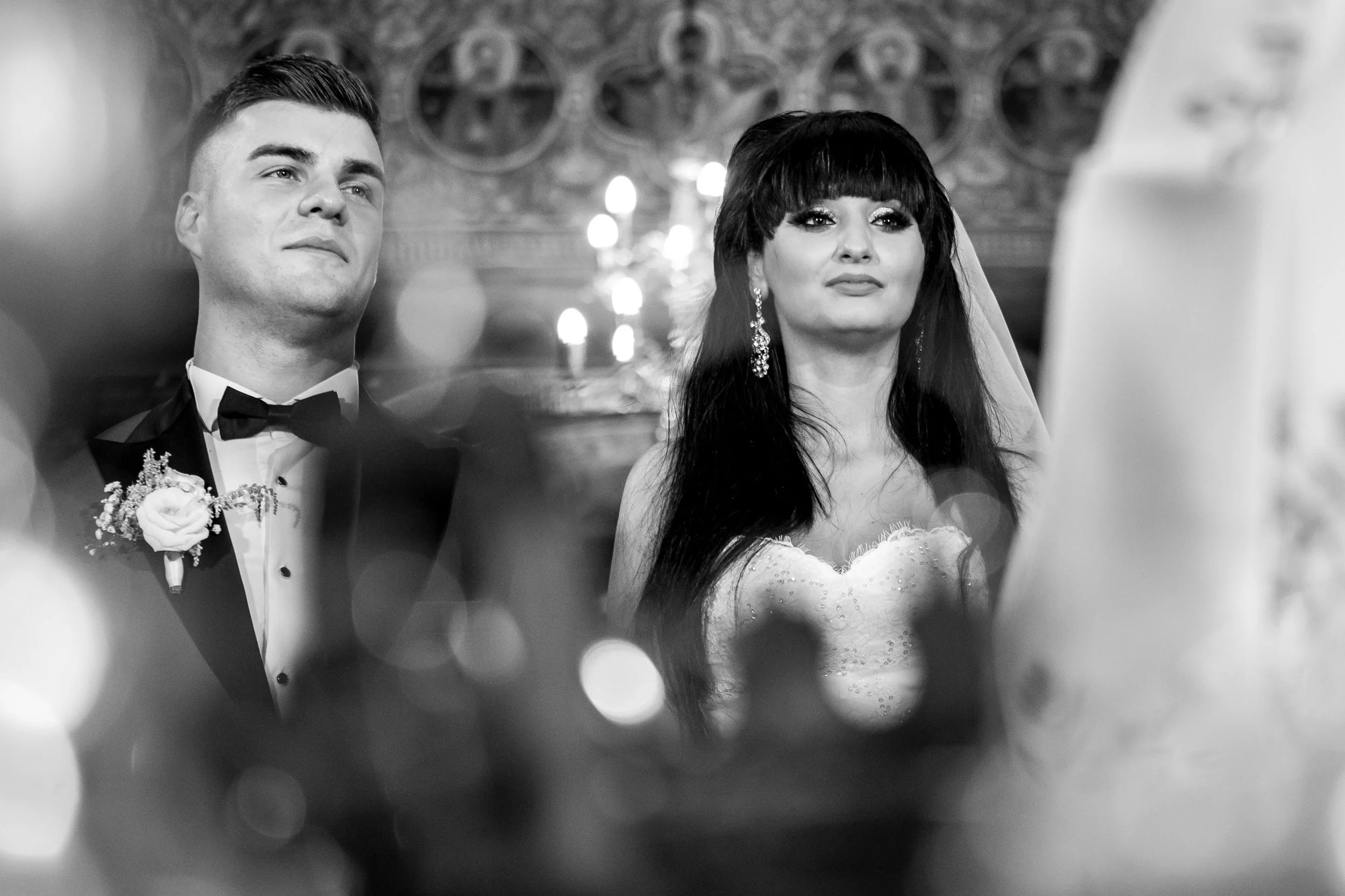 Black and white photo of a bride and groom during a wedding ceremony, facing forward, with blurred decorative background and circular light reflections.