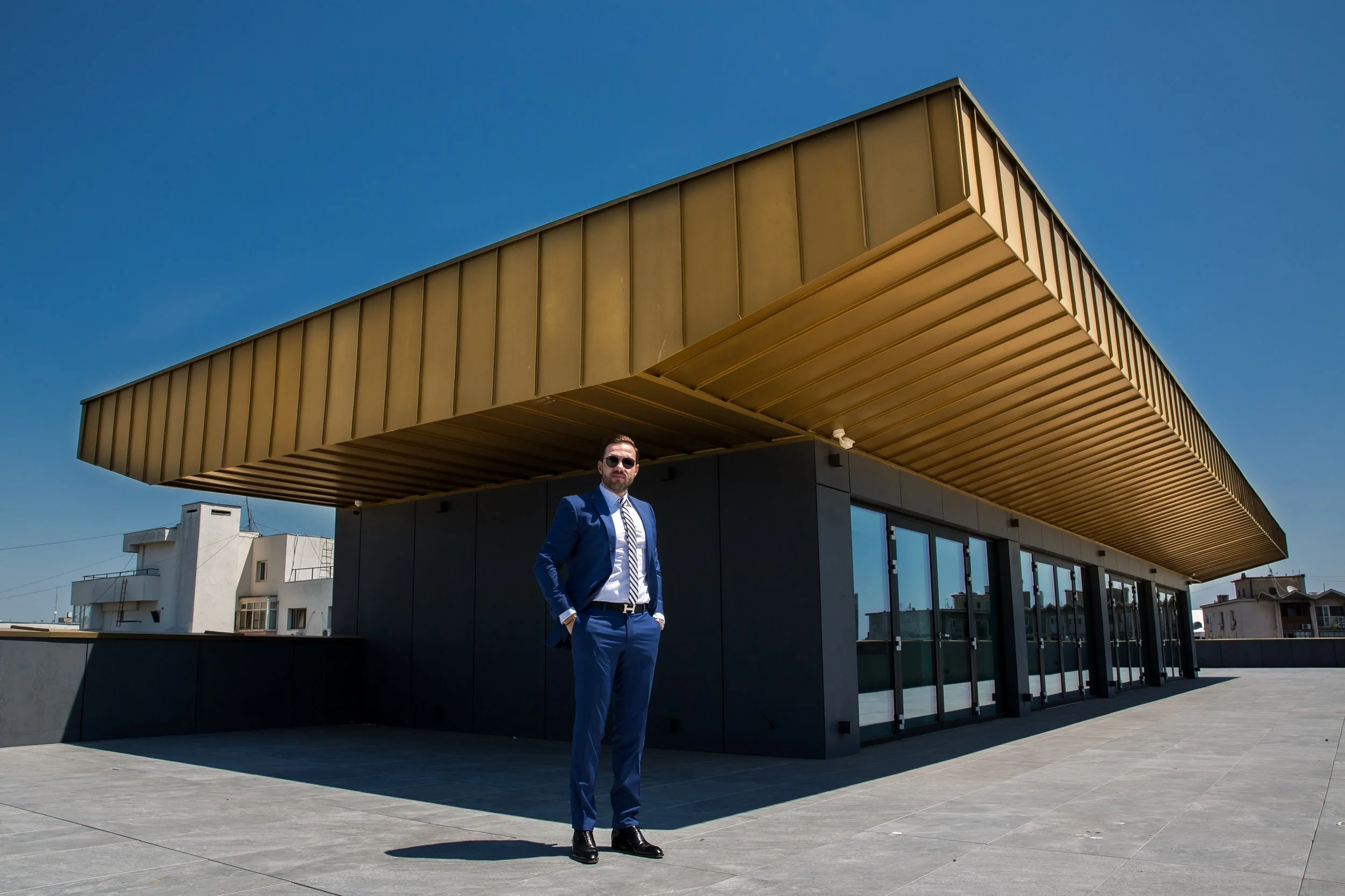 A man in a blue suit, white shirt, and striped tie standing on a modern rooftop terrace with a gold-colored architectural feature and blue sky in the background.